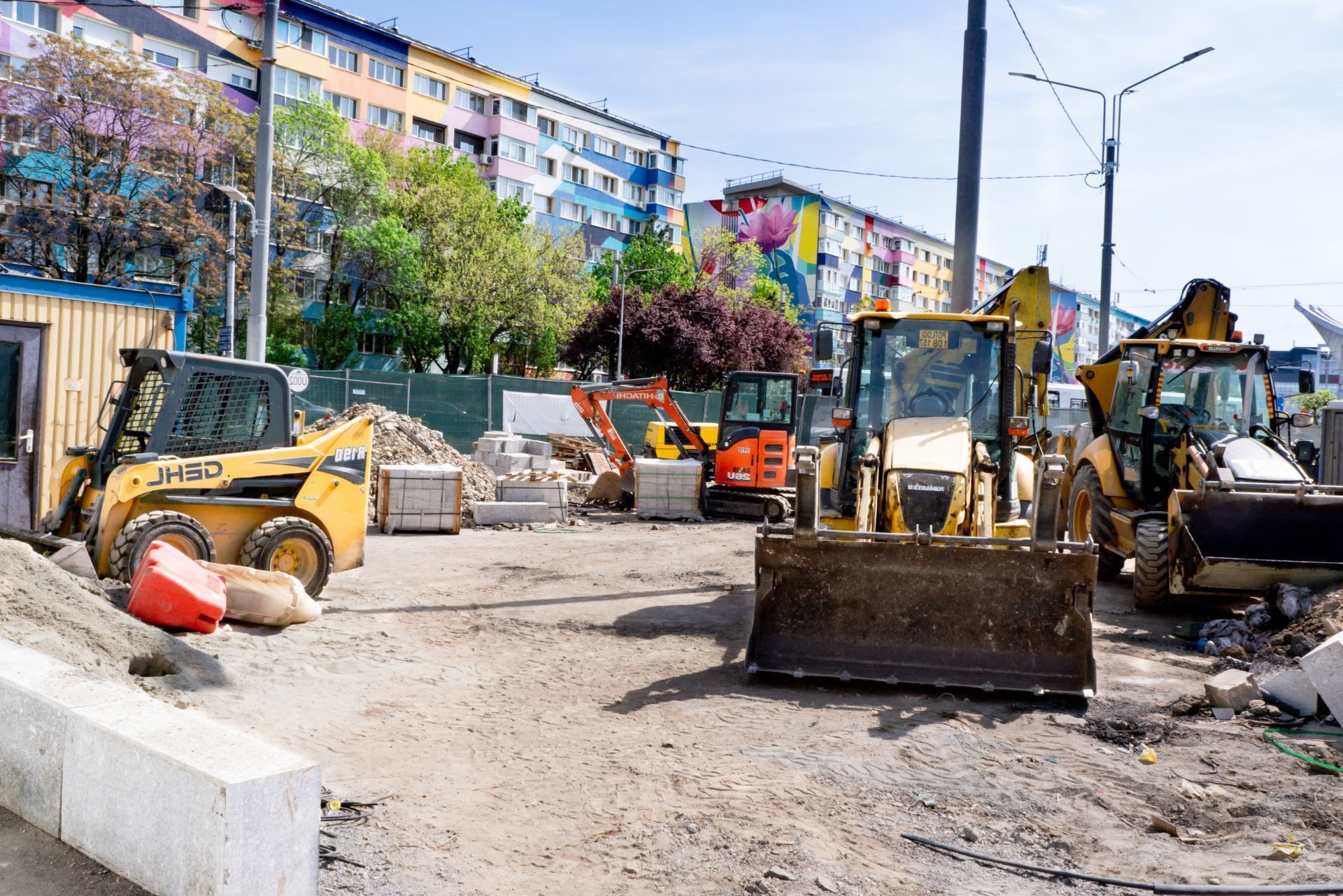 Construction site with yellow and orange heavy machinery, including excavators and a skid steer, in front of buildings.