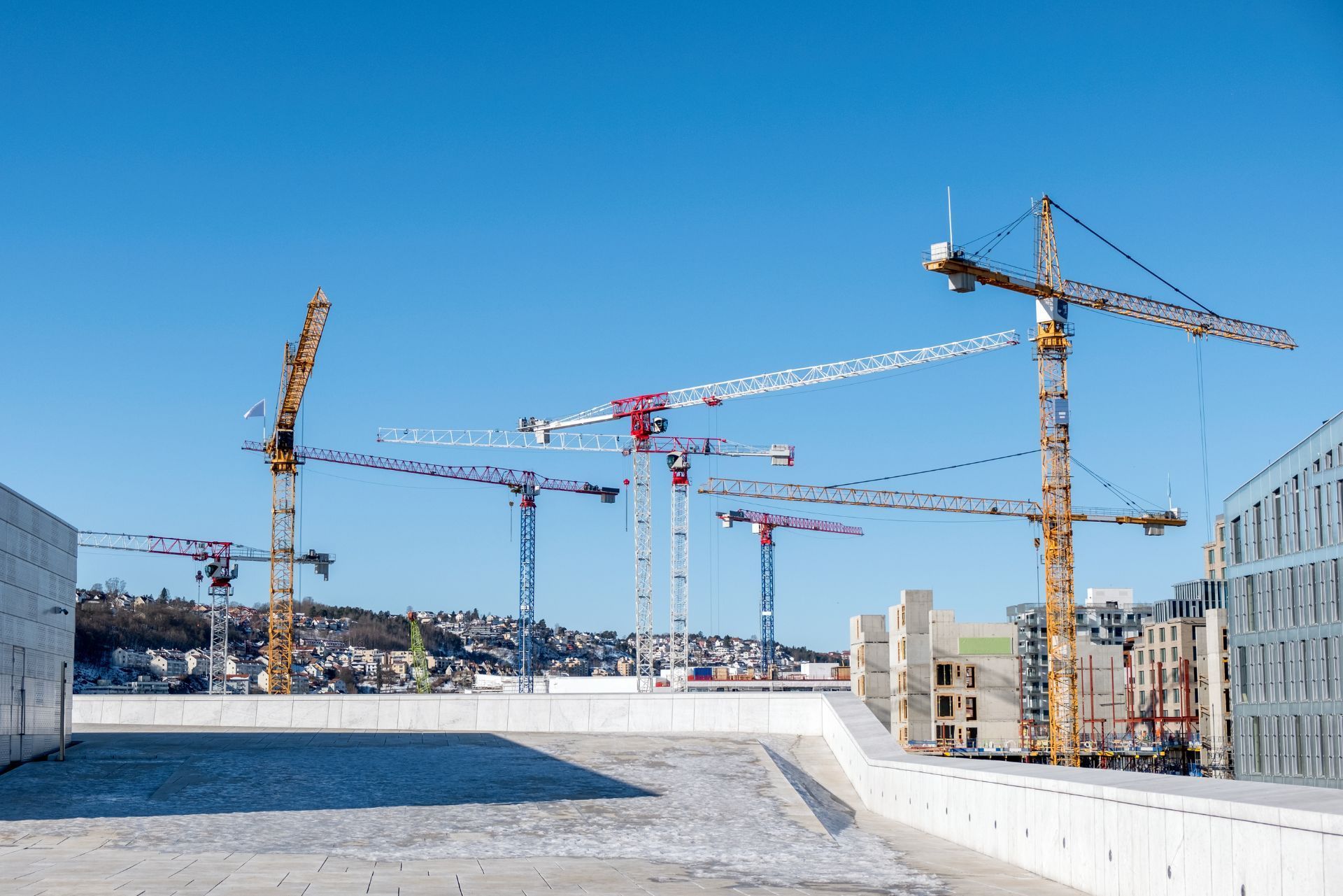 Multiple tall construction cranes tower over a construction site against a clear blue sky.
