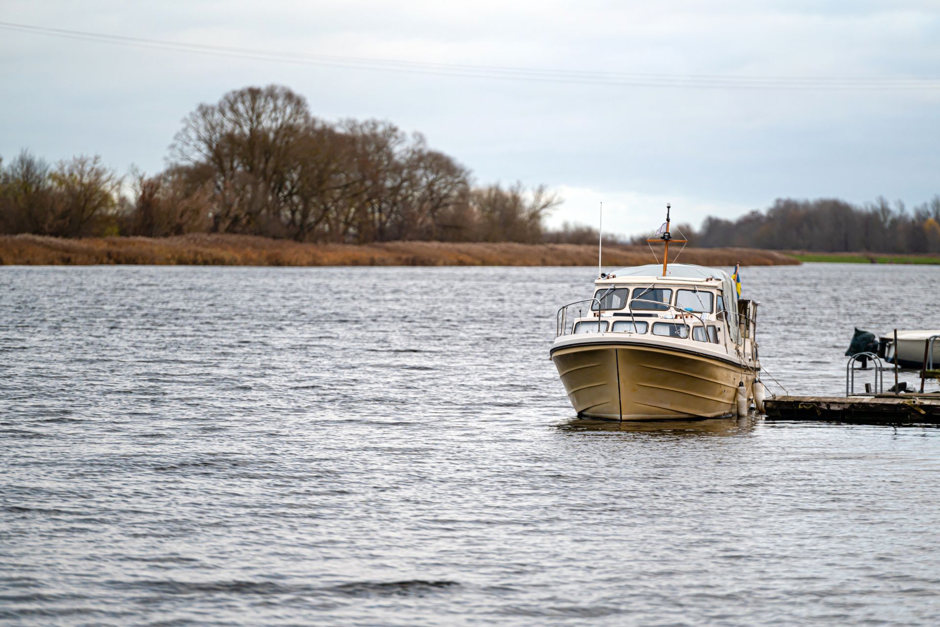Small tan boat docked on a gray river with bare trees along the opposite shore