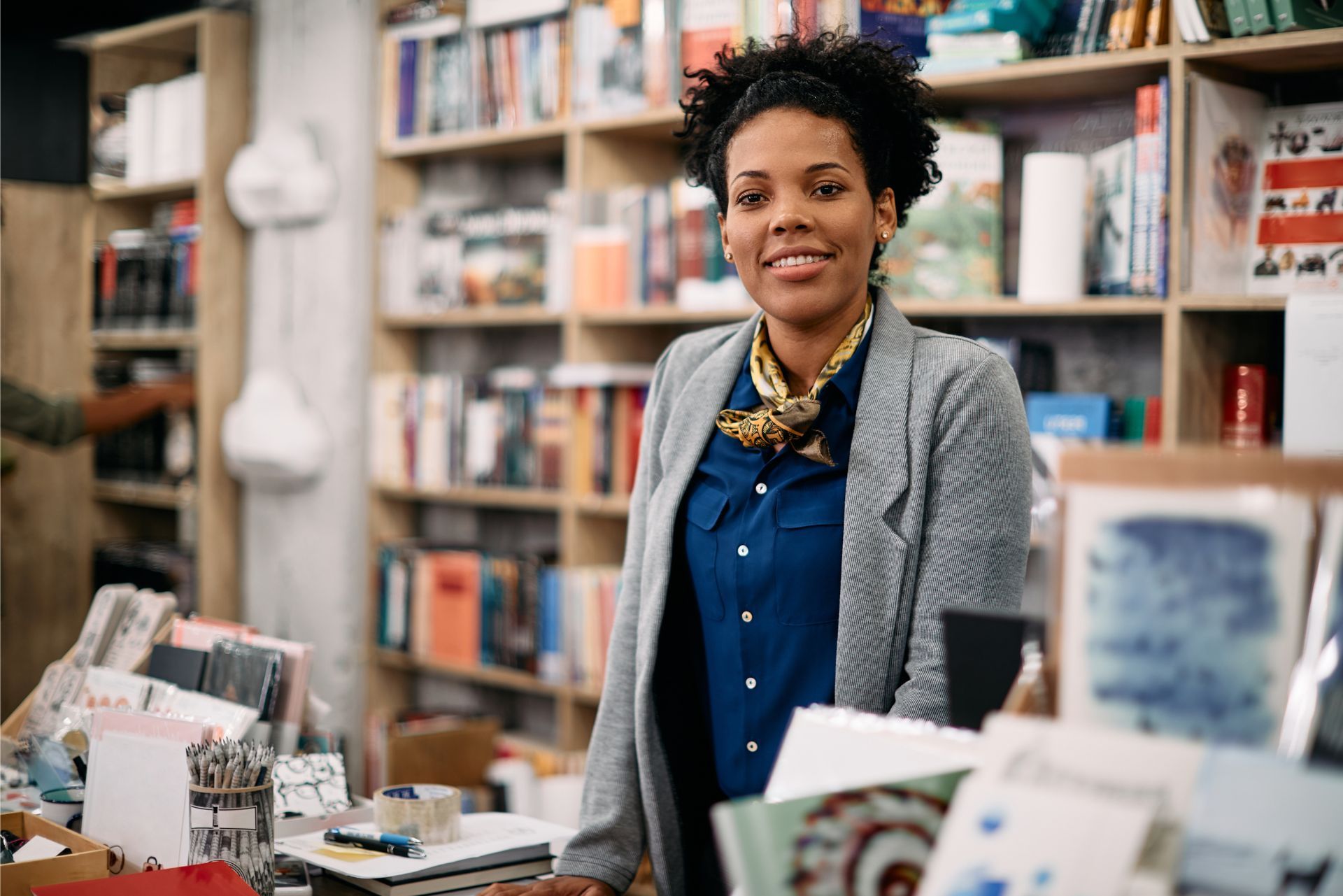 A person wearing a blue shirt and gray blazer smiles while standing in a bookstore filled with shelves of books.