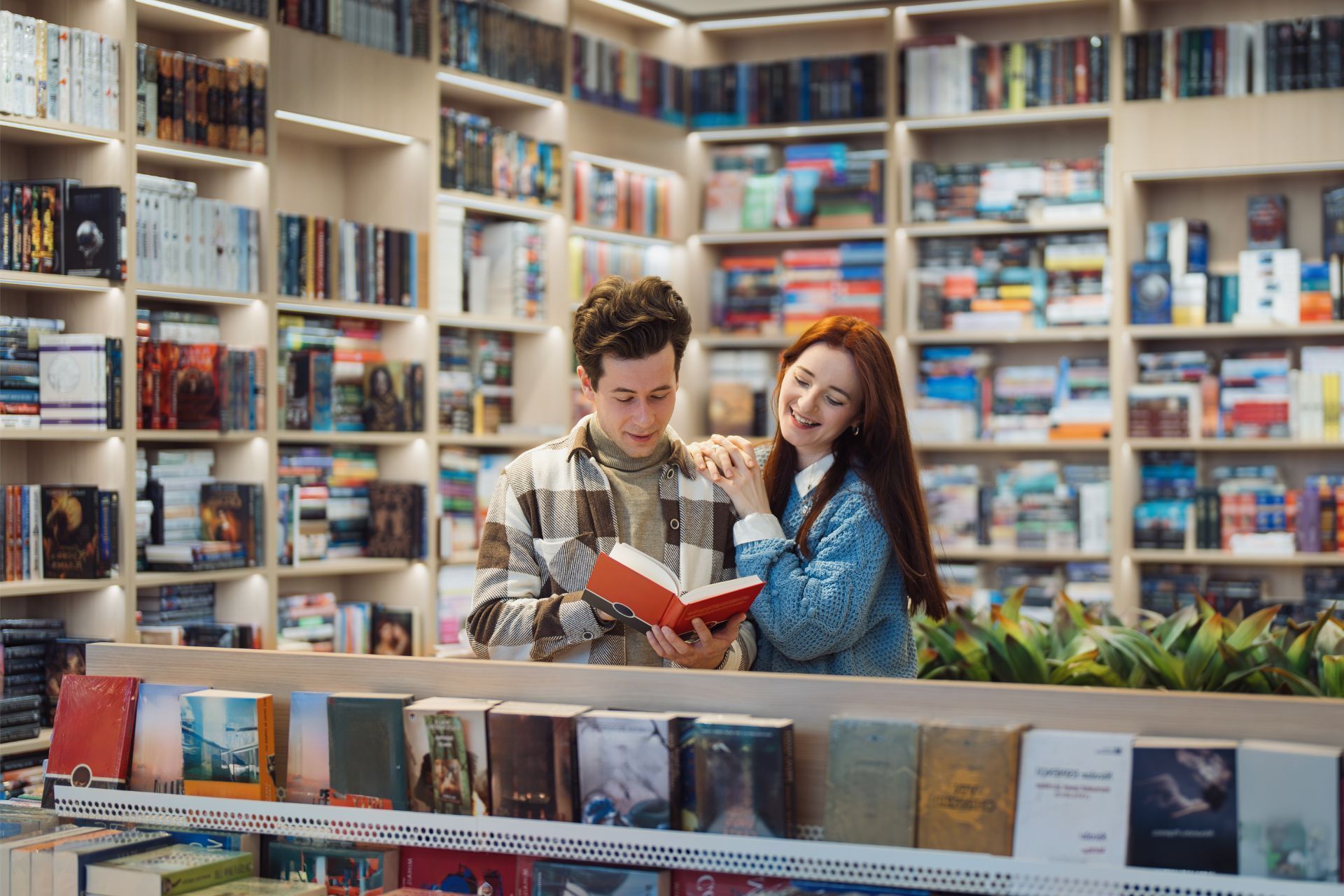 Two people reading a book together in a brightly lit bookstore surrounded by shelves filled with many books.