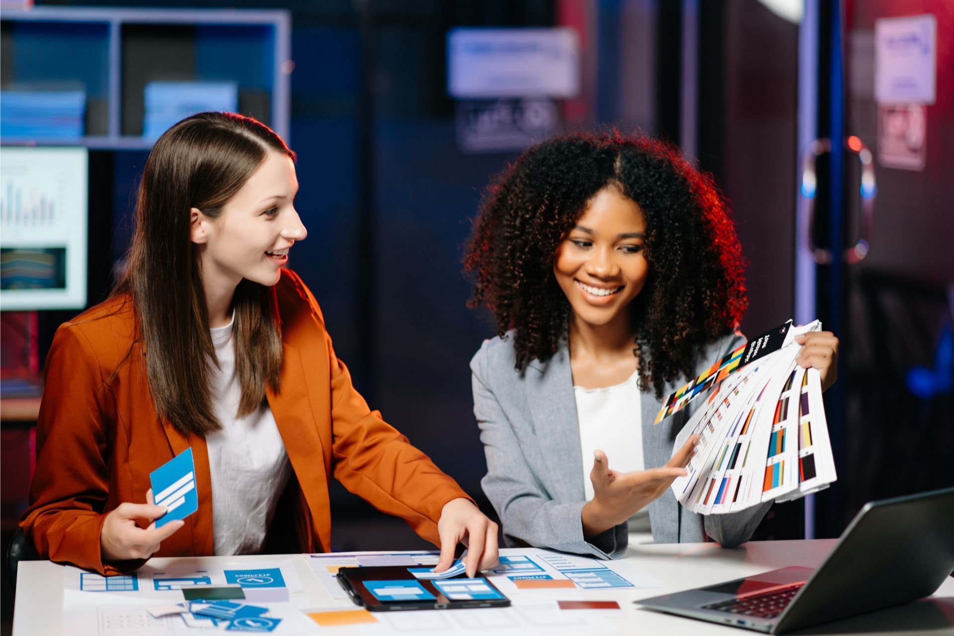 Two women reviewing design swatches at a desk with a laptop in a modern office