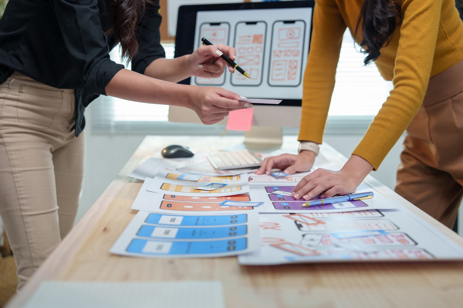 Two people reviewing a colorful chart on a table, one pointing while holding a pink cup, in a bright workspace