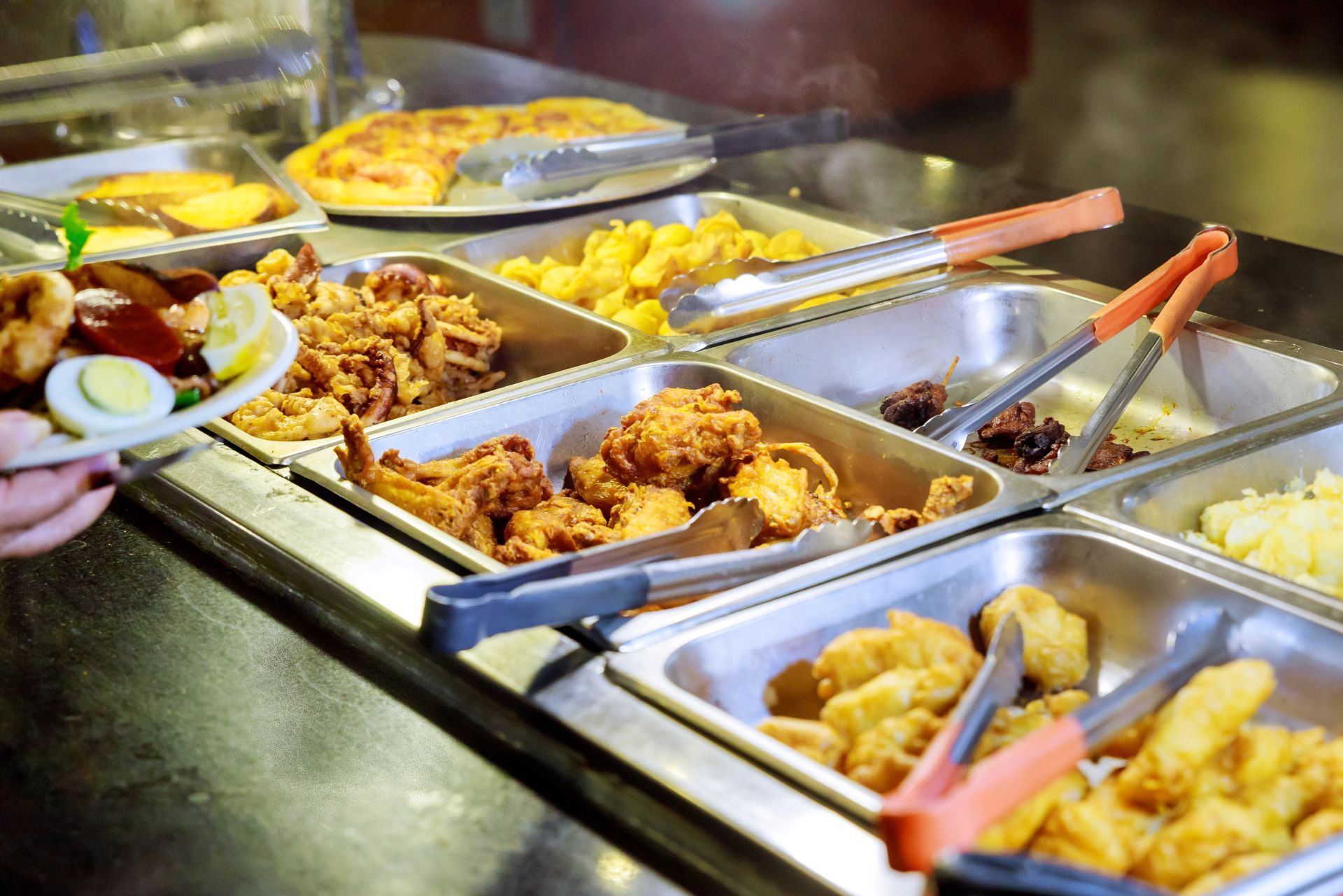 Buffet counter with trays of fried food, pasta, and vegetables; a hand serves a plate with salad.