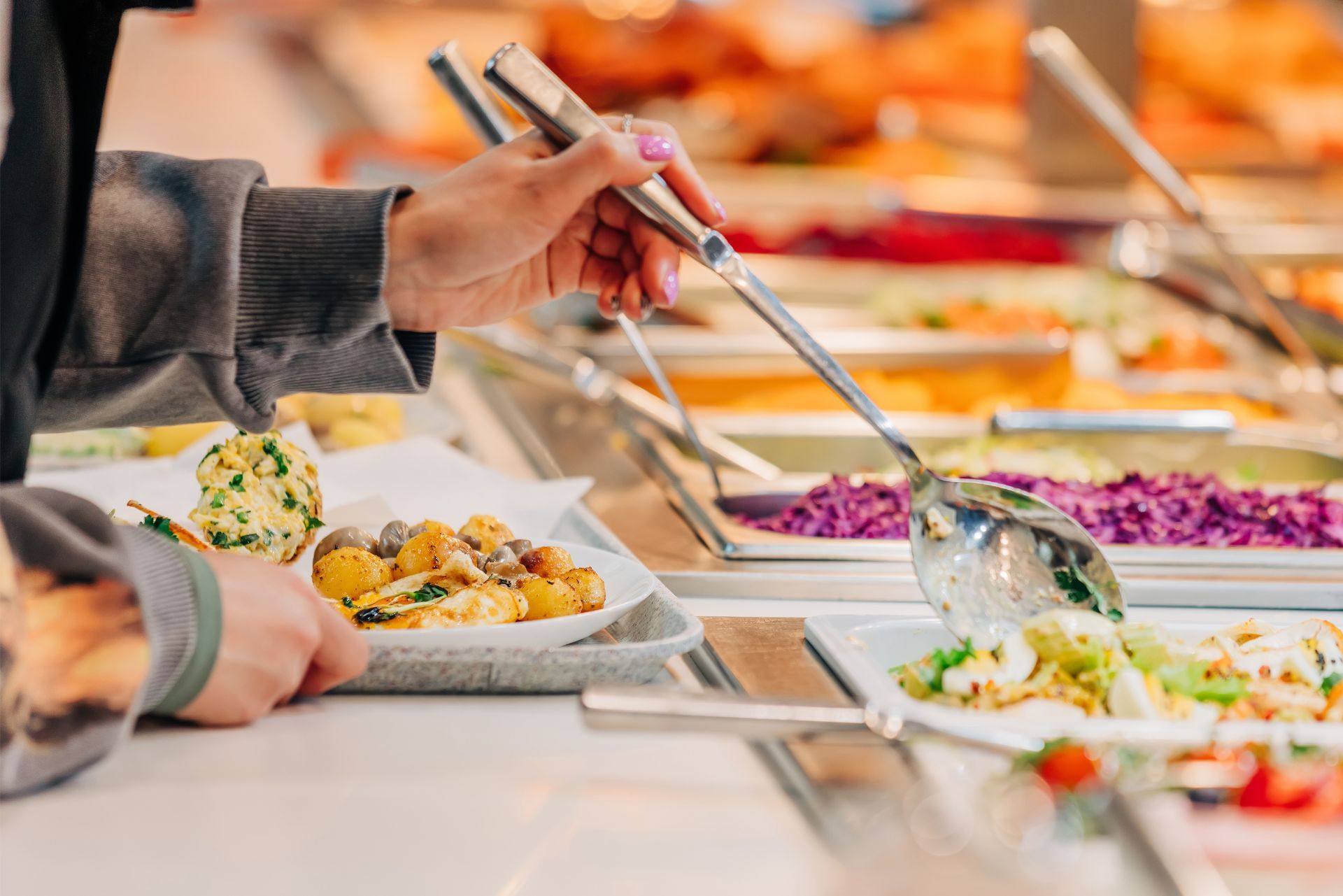 People serving themselves food at a buffet with trays of colorful dishes and serving tongs