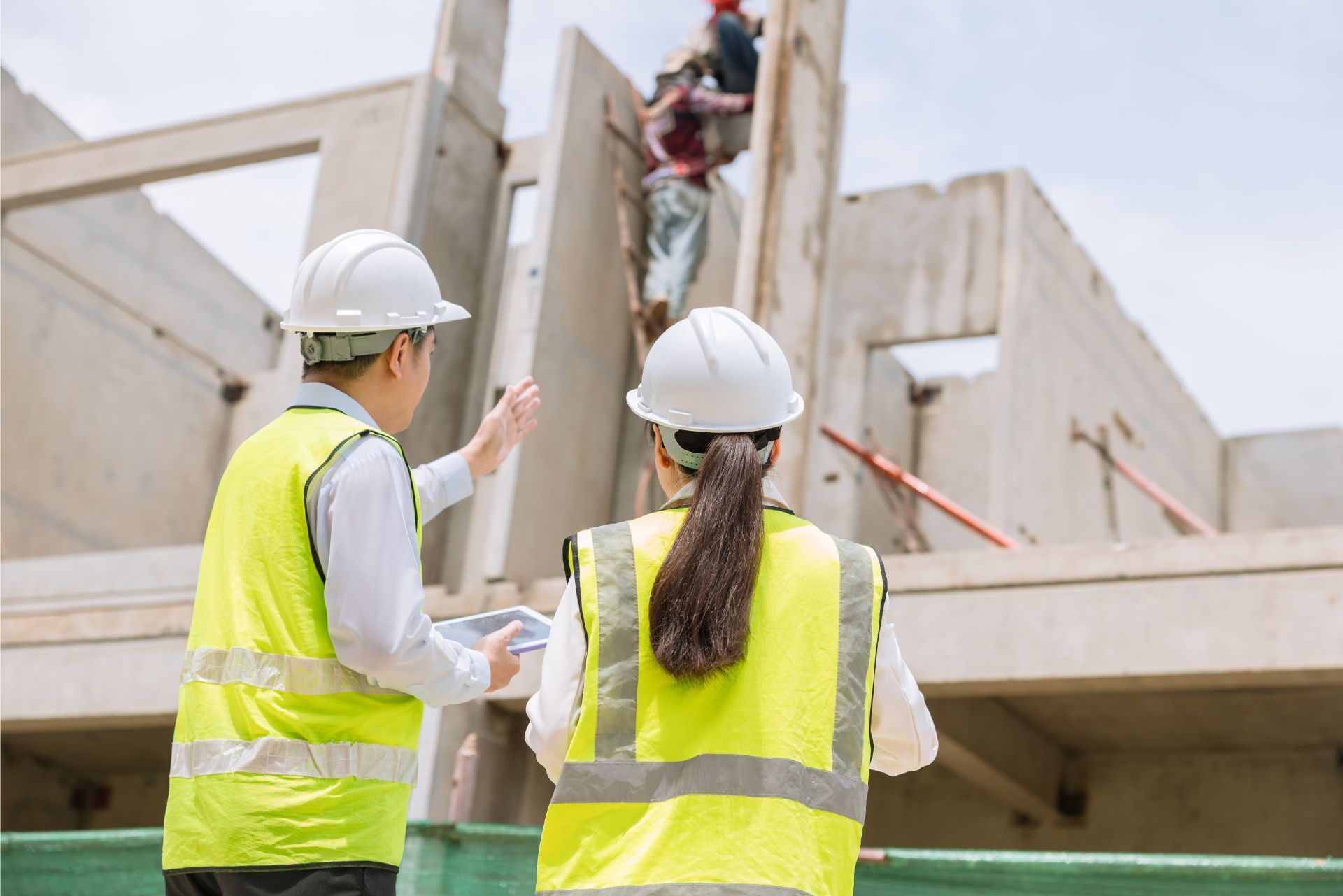 Two construction workers in hard hats and yellow vests inspect a building site while a worker climbs scaffolding.