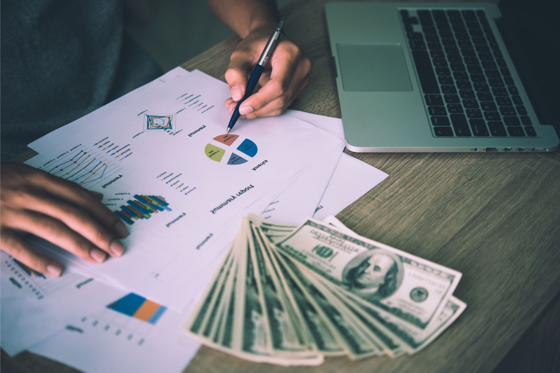 A person studies financial charts and graphs on paper while sitting at a desk with a laptop and a fan of US dollar bills.