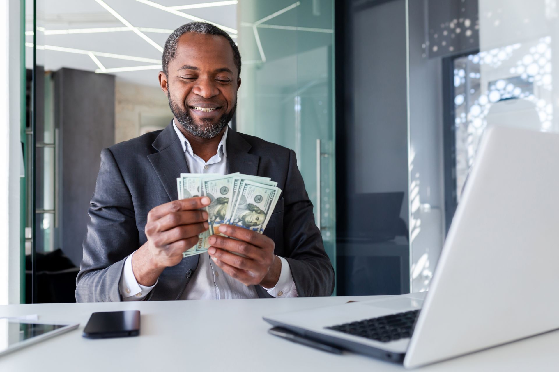 A professional sitting at a desk with a laptop, smiling while counting a stack of cash.