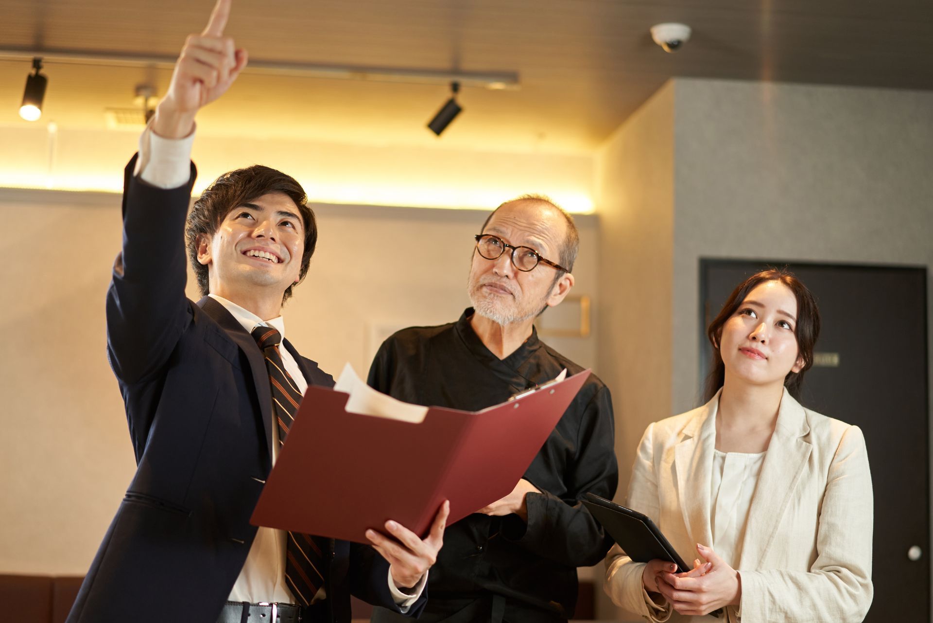 A man in a suit points to the ceiling while showing a red folder to two colleagues in an office setting.