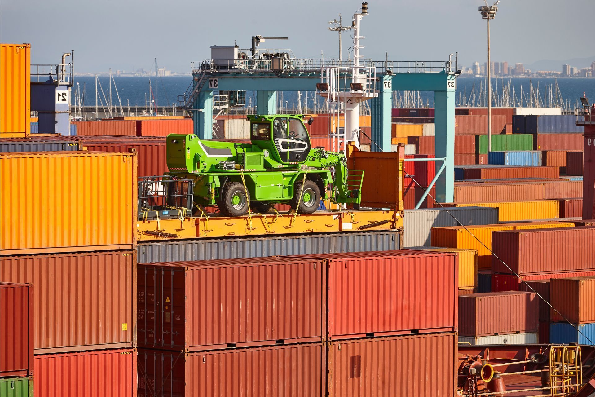 Green container handler in a busy shipping port with stacked orange cargo containers and cranes in the background