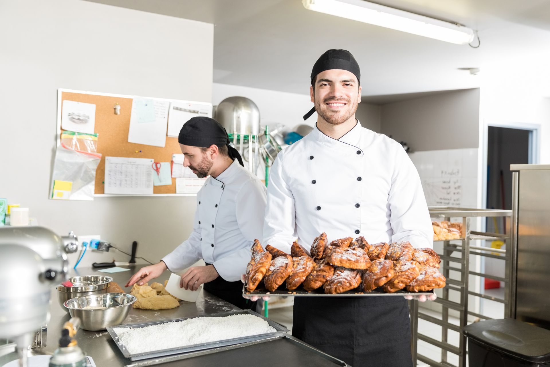 Two chefs in a bright kitchen, one holding a tray of baked pastries and the other preparing dough.