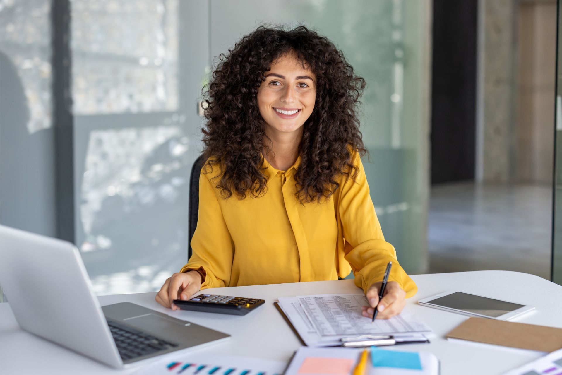 Woman in a yellow blouse smiling at a desk with a laptop, papers, and office supplies