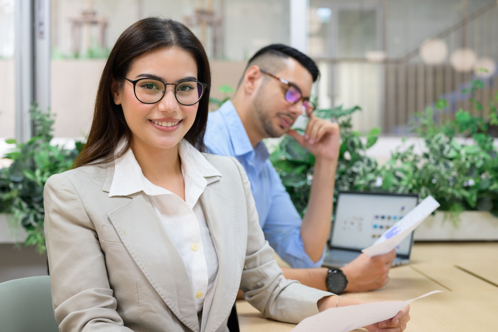 Smiling woman in beige blazer at desk, with man working on laptop in the background
