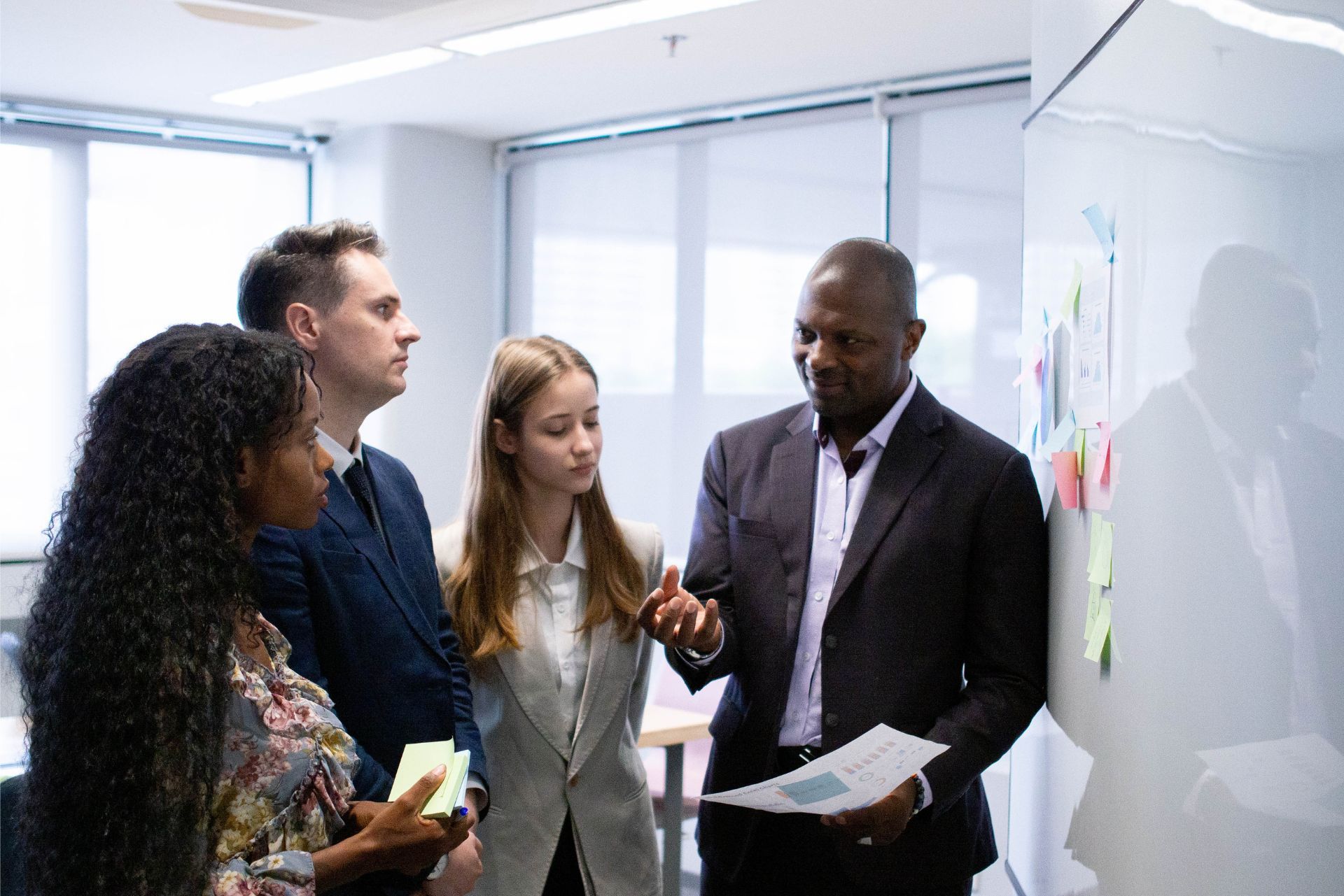 Four coworkers discussing notes beside a whiteboard in a bright office meeting room