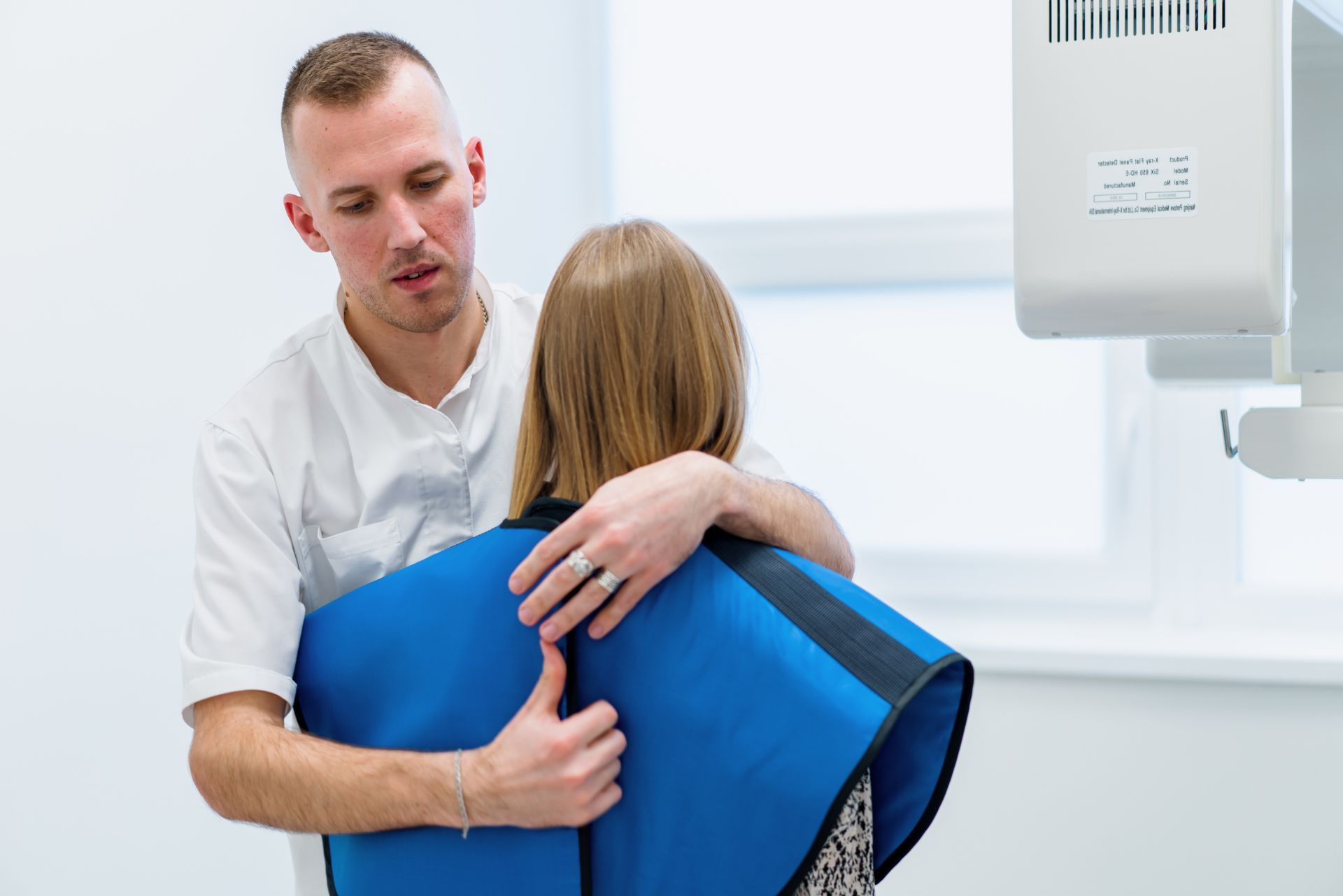 A medical professional in a white uniform secures a blue radiation protection cape around a patient before an X-ray.