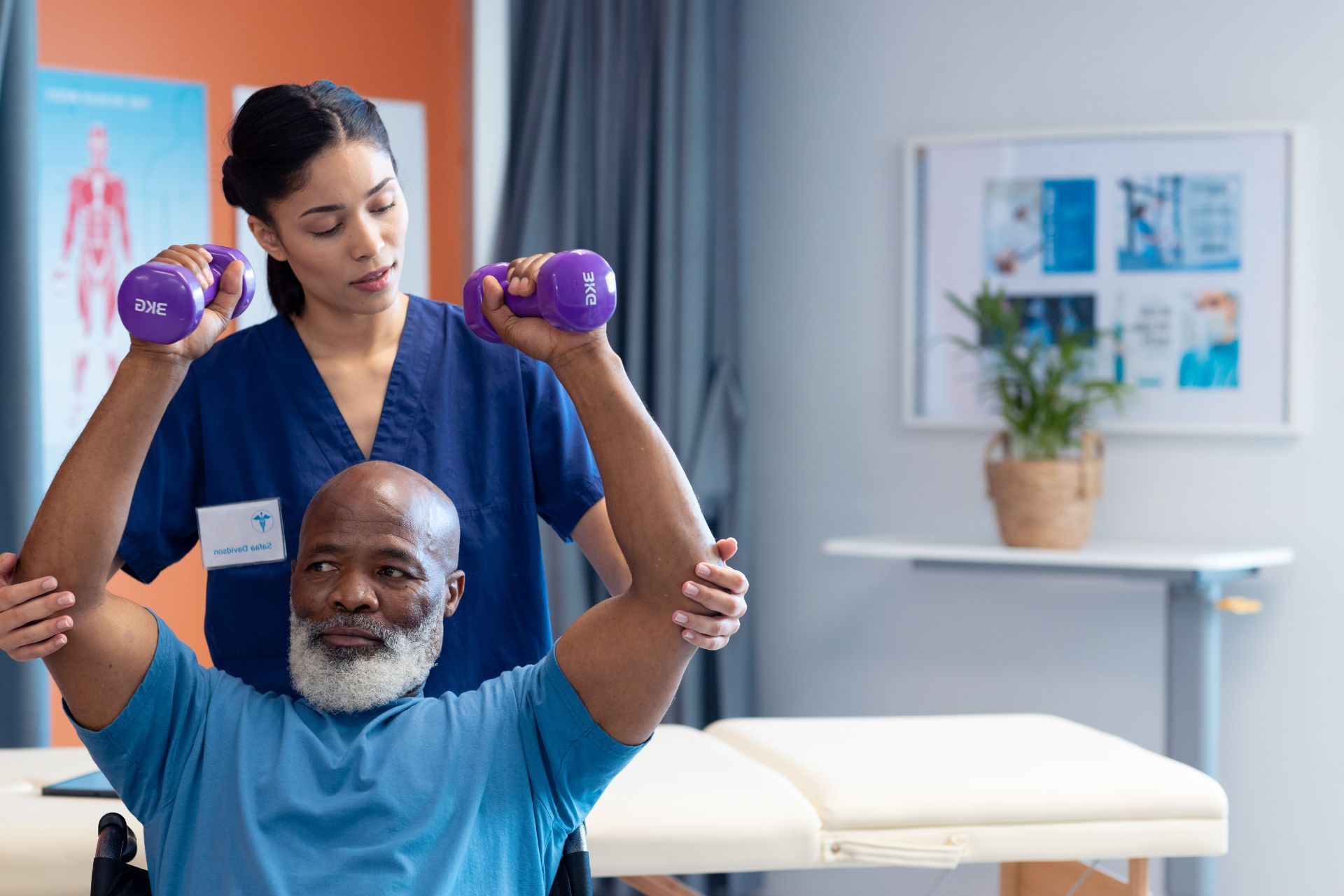 A healthcare professional assists a patient in a blue shirt with arm exercises using small purple dumbbells.