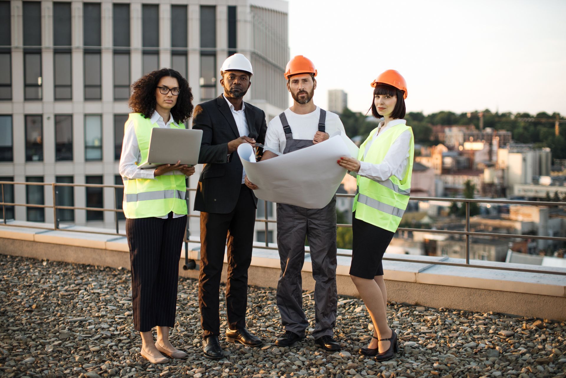 Four construction professionals in safety vests and hard hats reviewing blueprints on a rooftop.