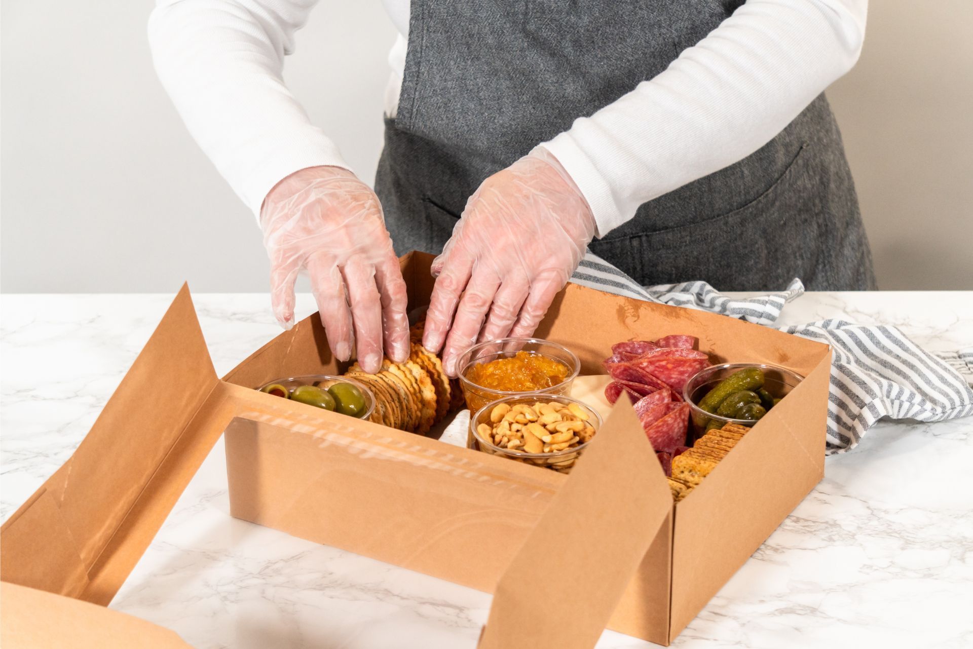 Hands arranging assorted baked goods in an open cardboard box on a marble table