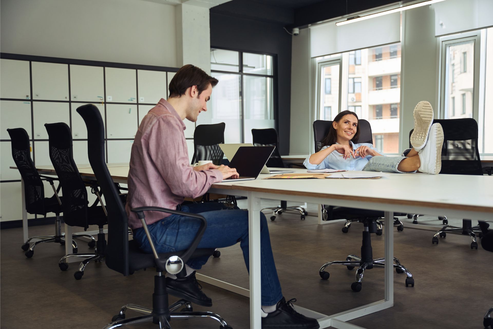 People working in a modern office, one seated at a laptop and another gesturing across the table.