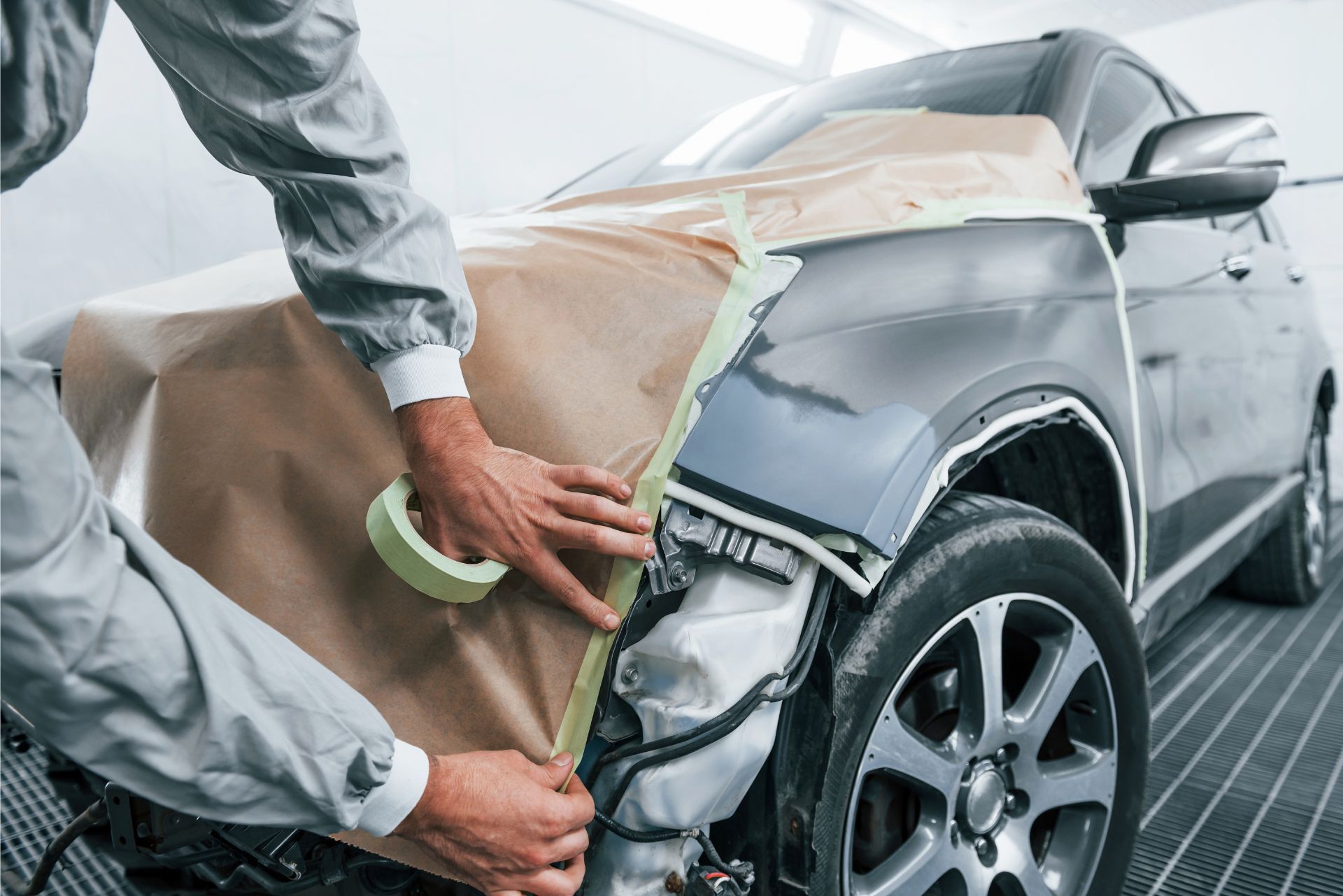 Person masking a damaged silver car’s front fender with brown paper and green tape in a repair shop