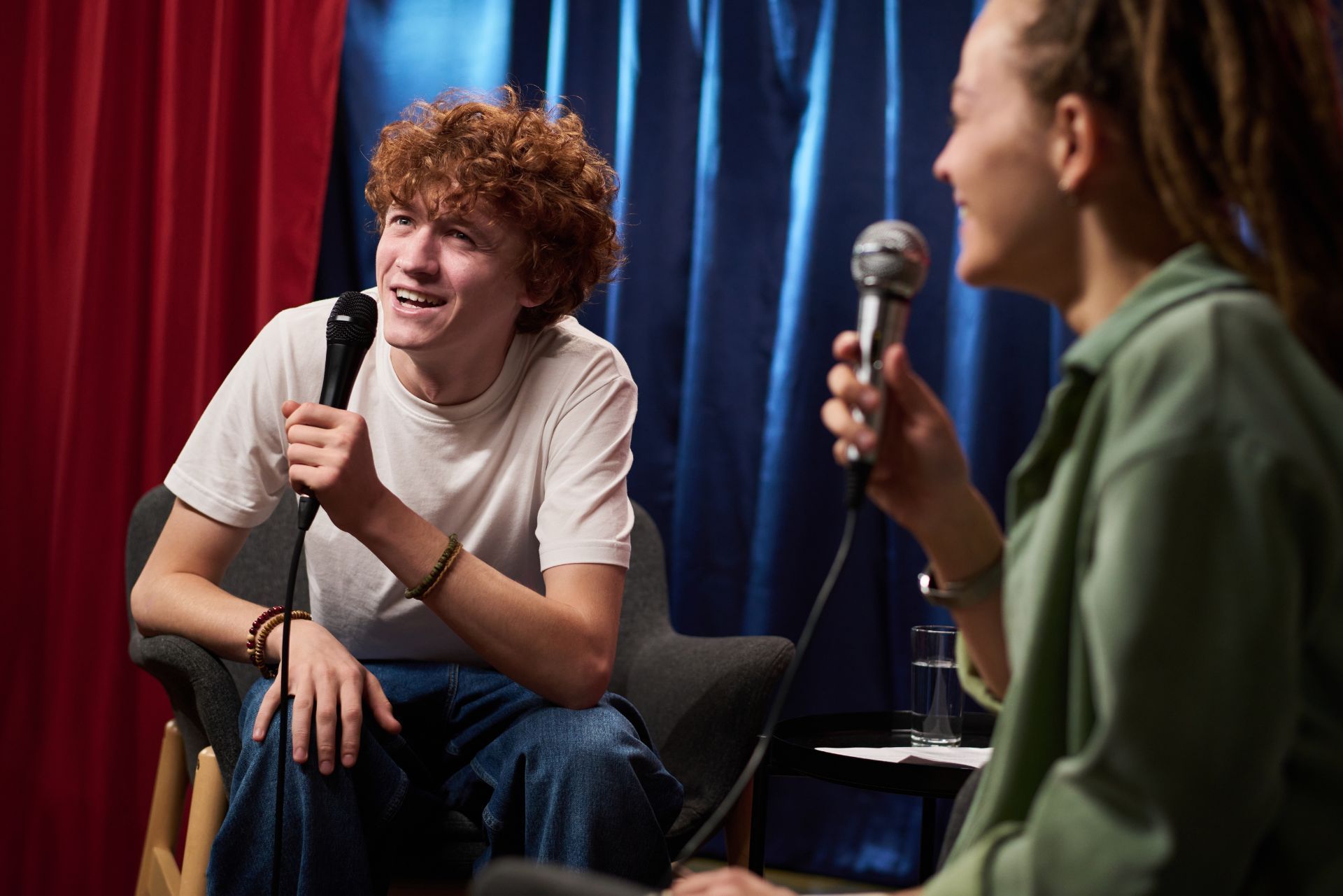 Two people holding microphones in a colorful stage setting, one smiling while seated near red and blue curtains