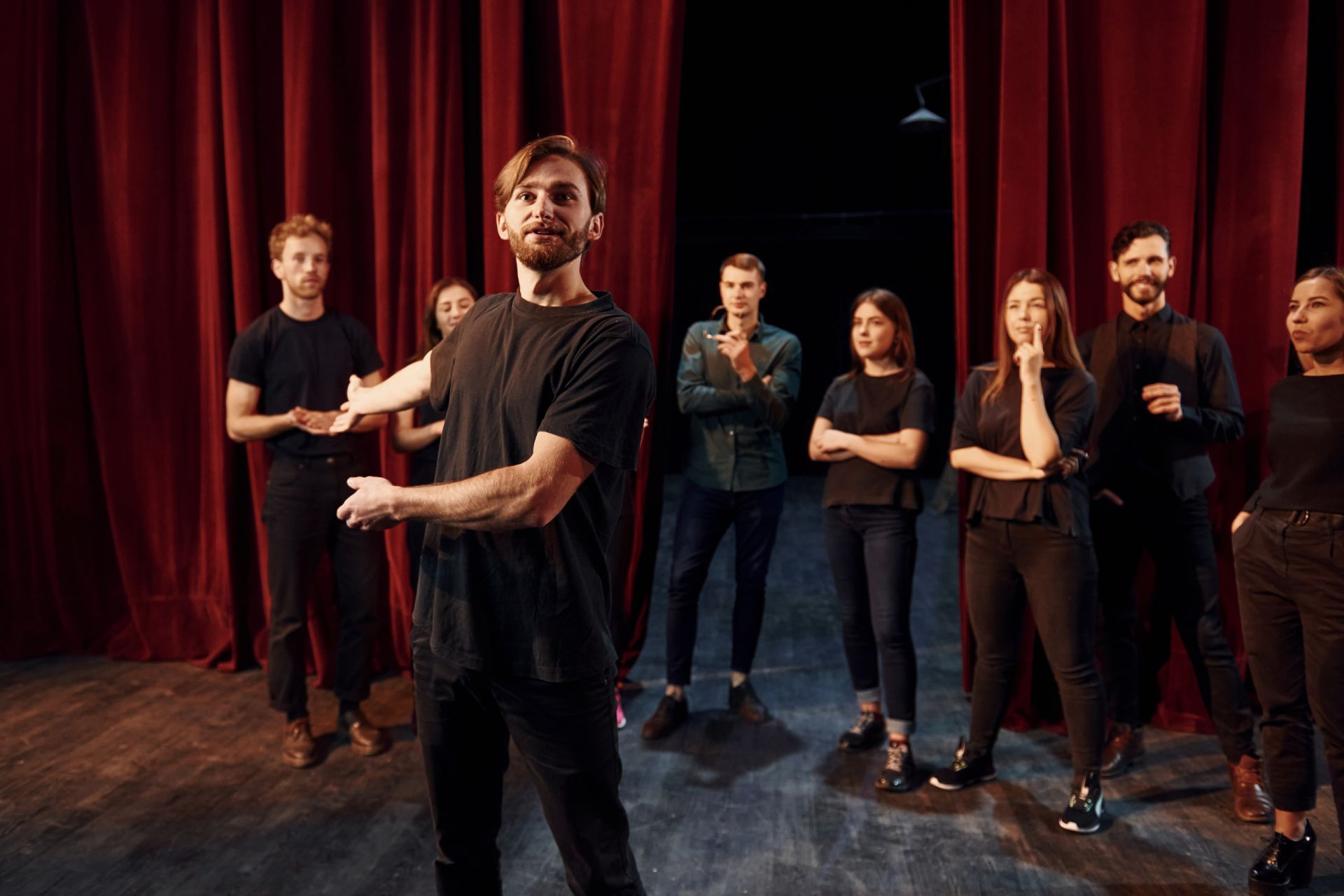 A group of performers stands on a stage with red curtains, one man gesturing in front while others watch.