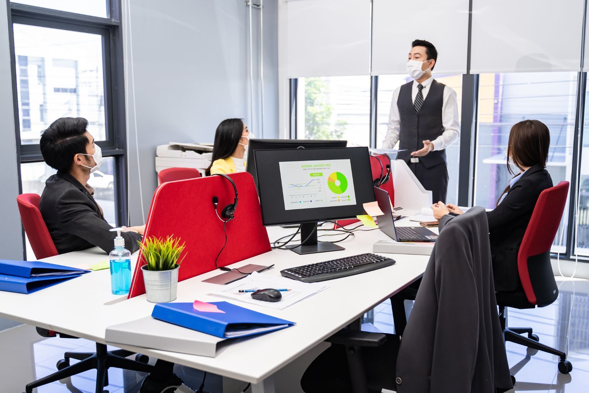 Business meeting in a bright conference room, with one presenter standing by a screen and three colleagues seated סביב a table