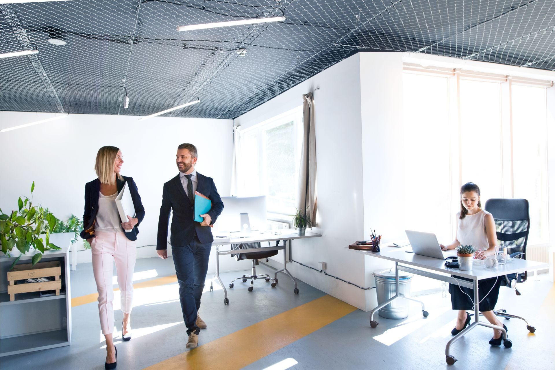 Two coworkers walking and talking in a bright modern office, with a seated person at a desk nearby.