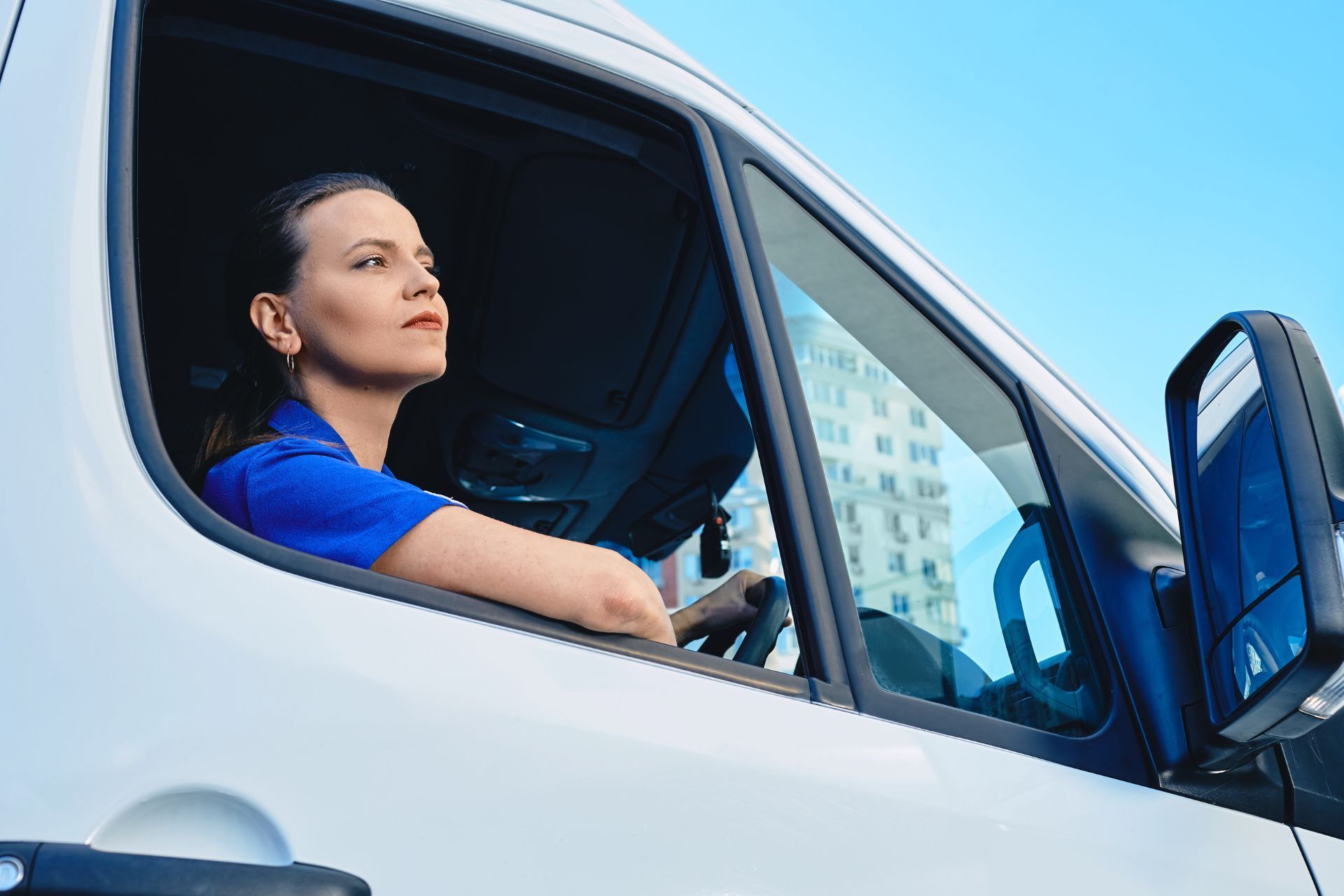 Woman driving a white van, seen through the open window on a sunny day.