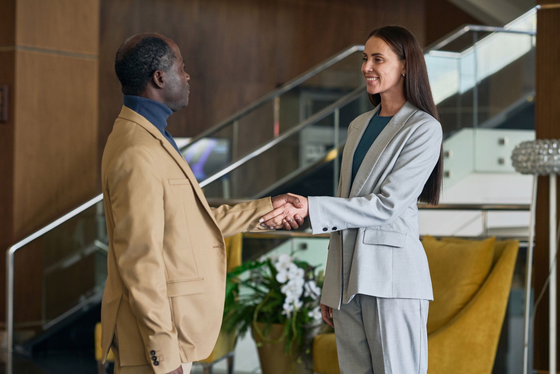 Two professionals shaking hands in a modern office lobby, smiling beside a staircase and plants.
