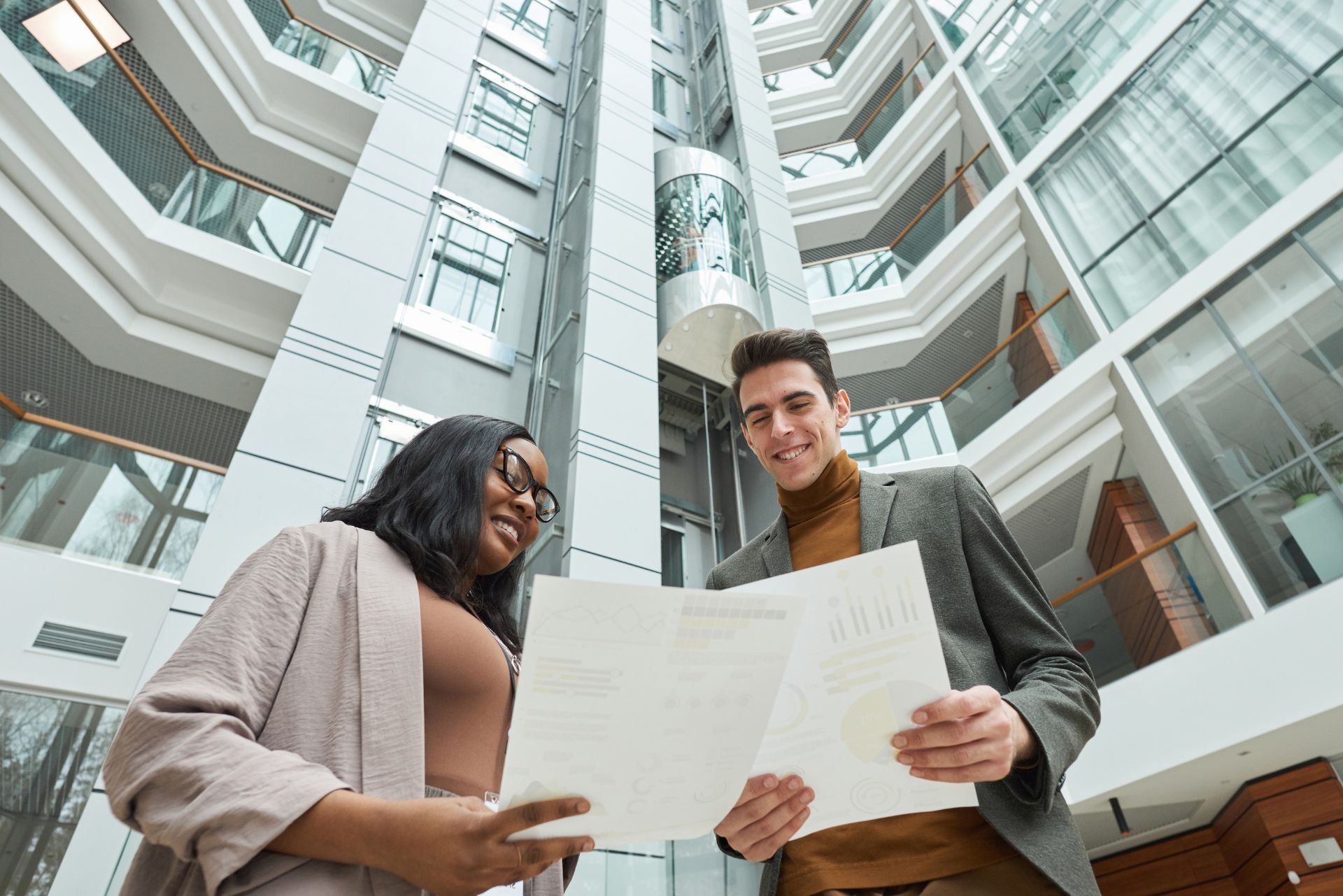 Two colleagues in business casual attire review documents together in a modern, multi-story building atrium.