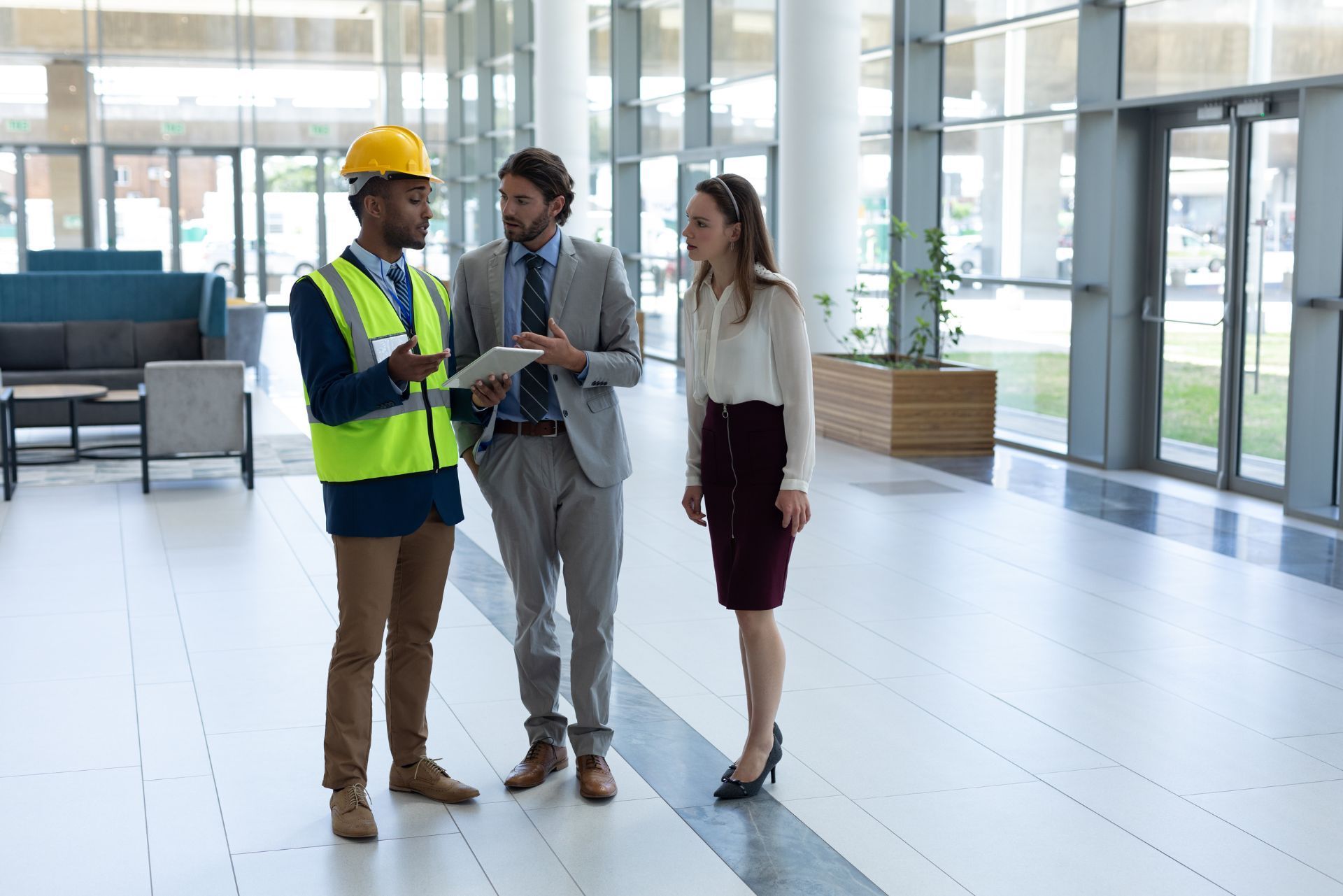 Three professionals in formal and high-visibility attire discuss a tablet inside a modern office lobby.