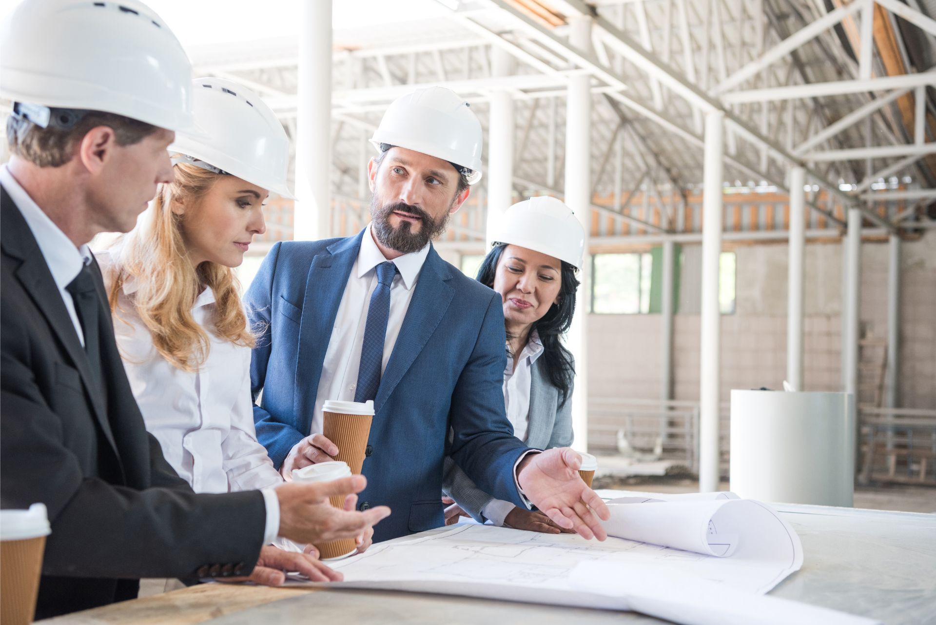 Engineers in hard hats review blueprints at a construction site, discussing plans around a table.