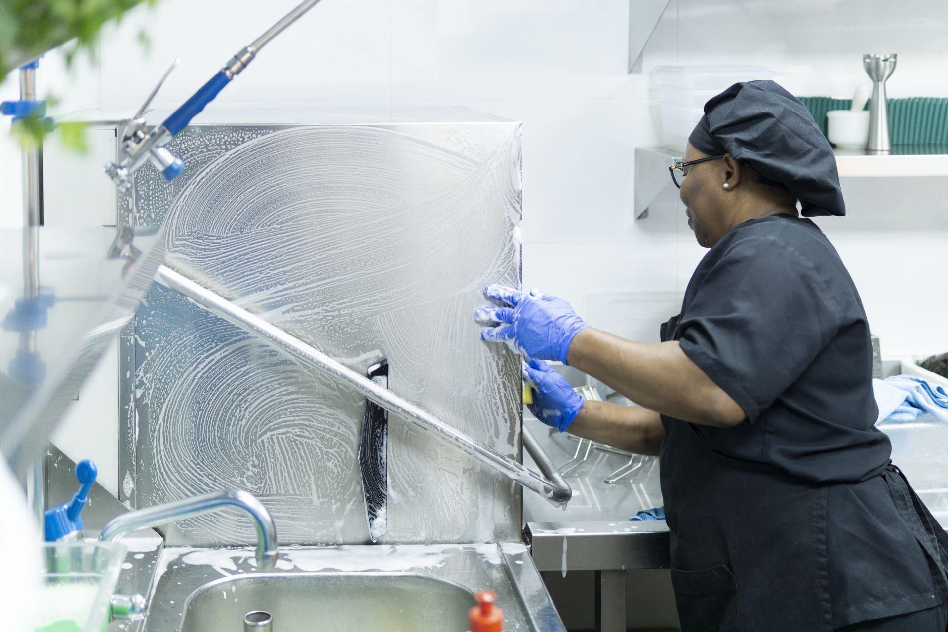 Worker in black uniform washing a large metal kitchen machine in a commercial kitchen