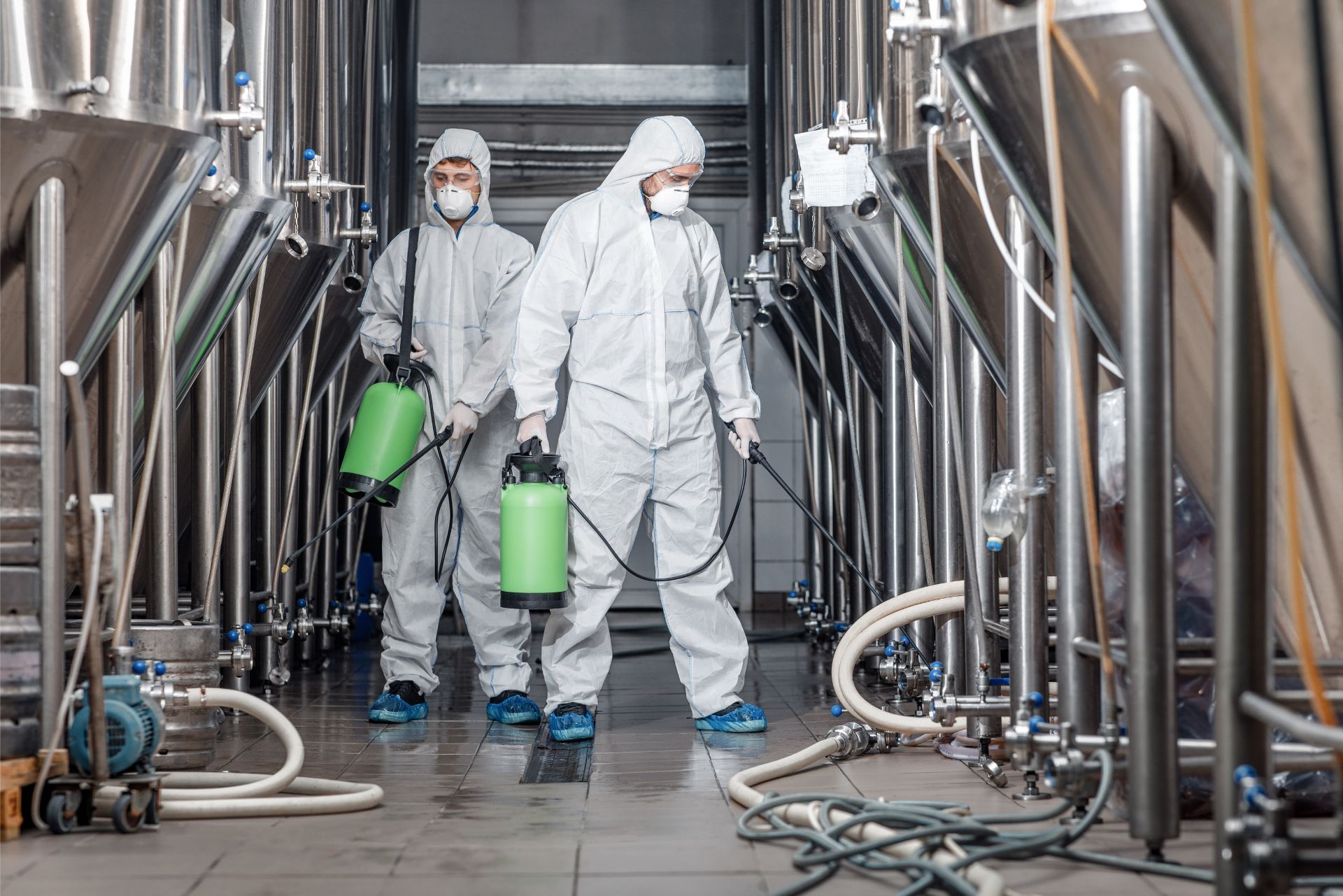 Two workers in white protective suits carry green canisters through an industrial facility with stainless steel tanks.