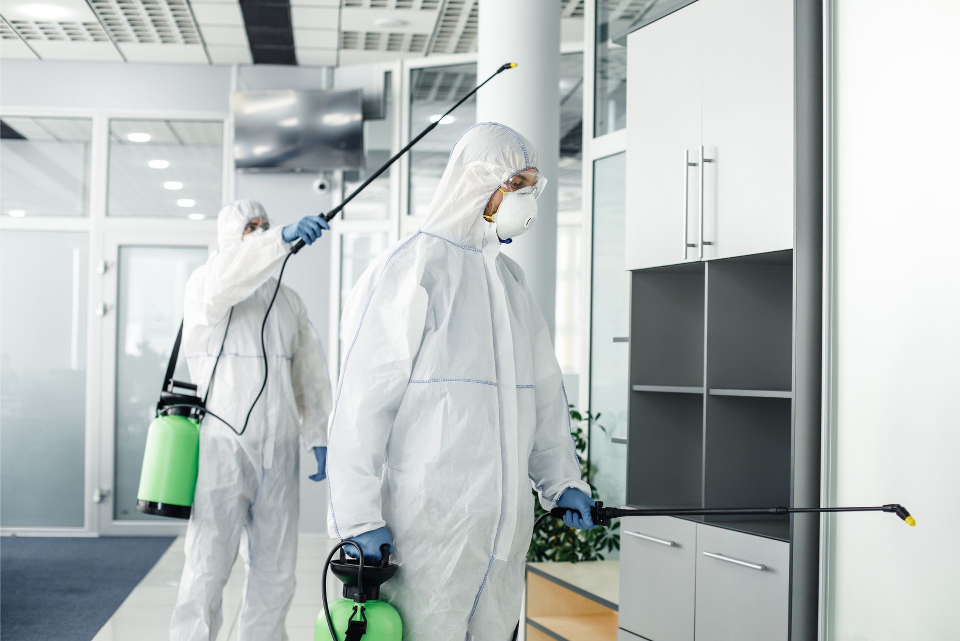 Two workers in white hazmat suits disinfecting a bright office with sprayers