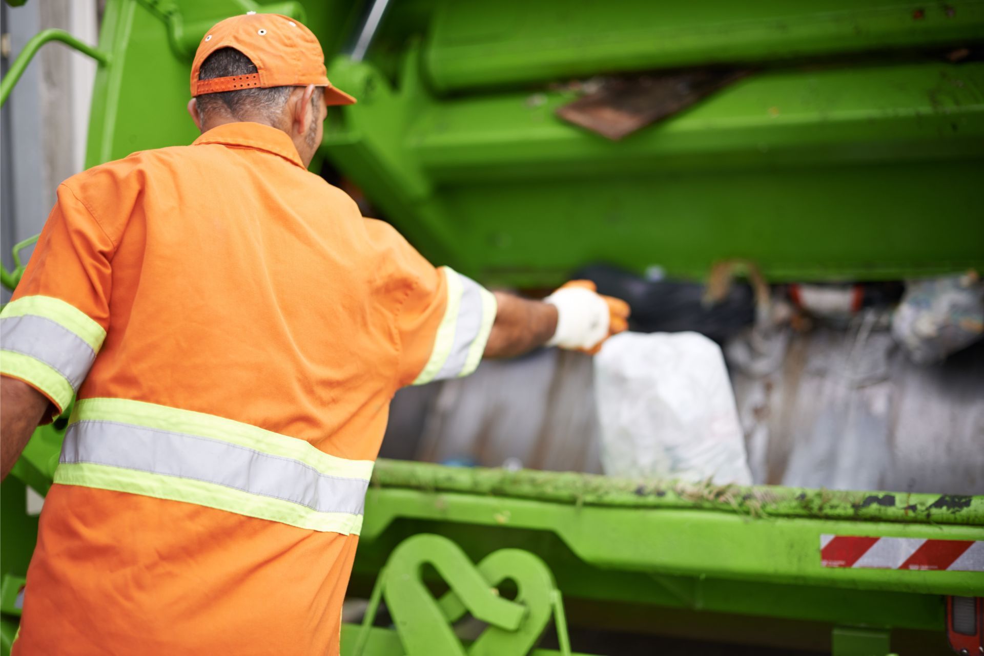 Worker in orange safety gear loading trash bags into a green garbage truck