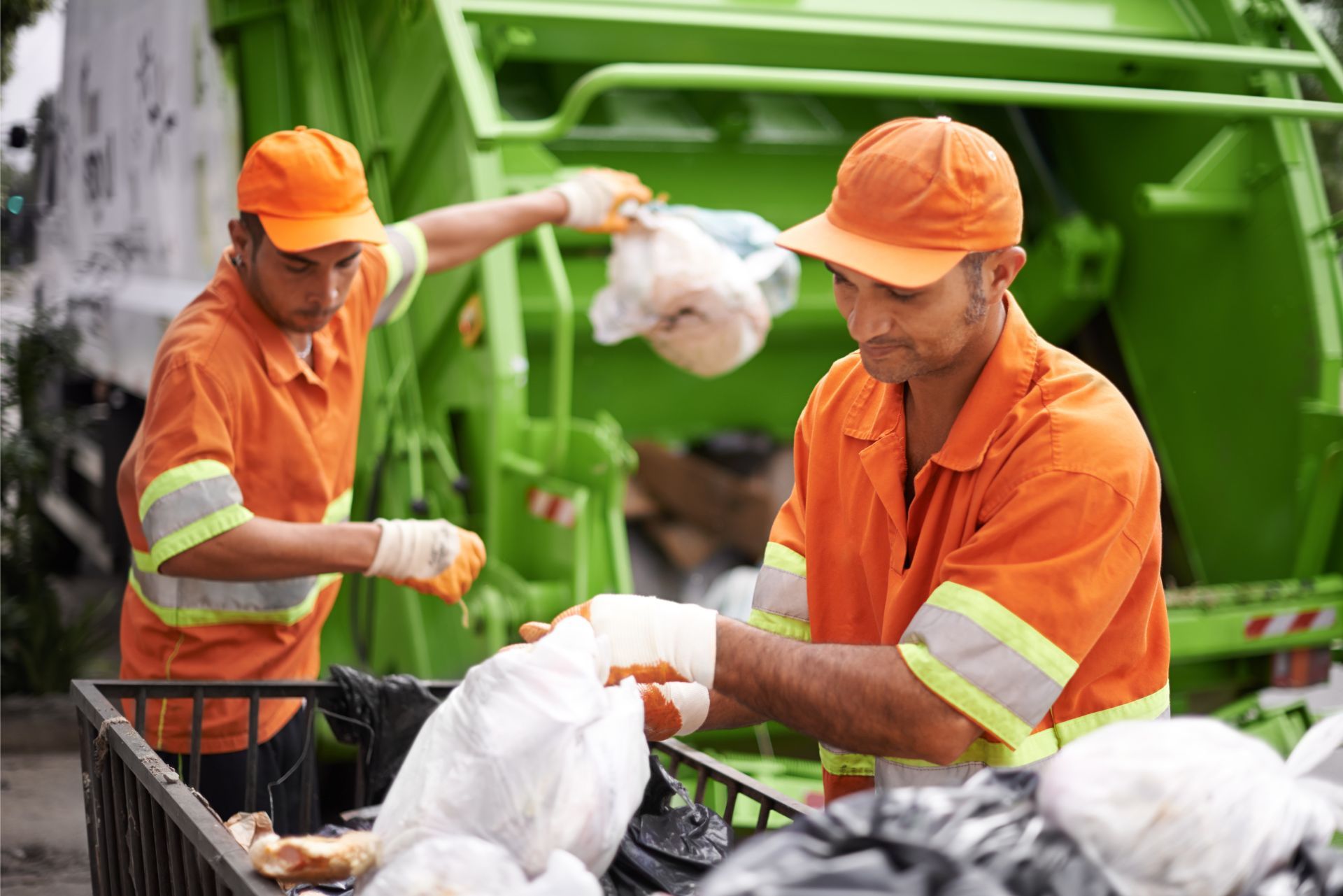 Two sanitation workers in orange uniforms sorting trash beside a green garbage truck