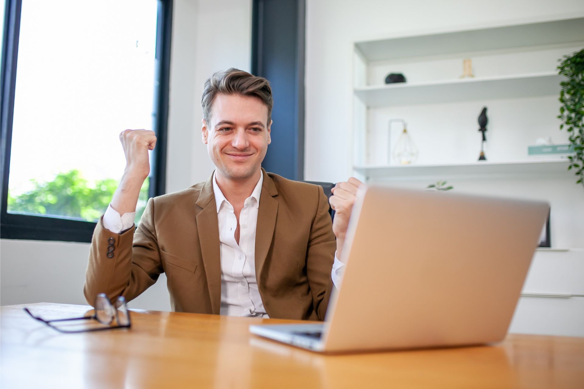 Smiling man in a brown blazer sits at a desk with a laptop in a bright office, raising one fist in celebration