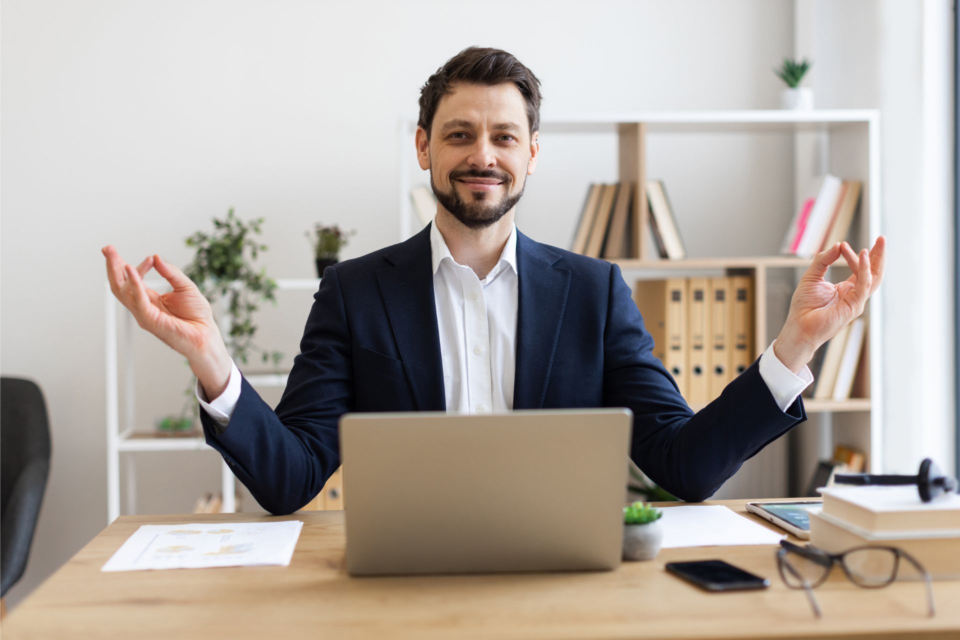 Smiling man in a navy blazer meditating at a desk with a laptop in a bright office