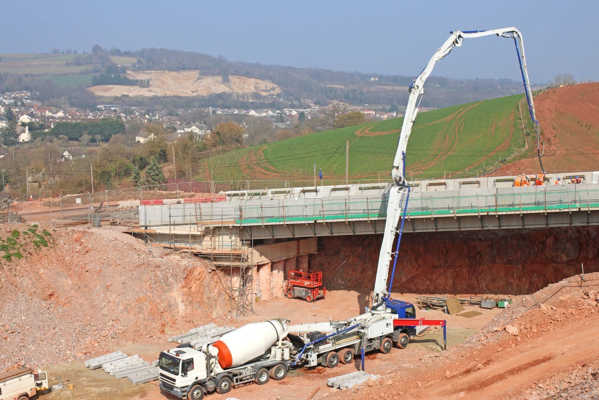 A concrete pump truck extends its boom to pour concrete onto a bridge construction site in a rural, hilly landscape.