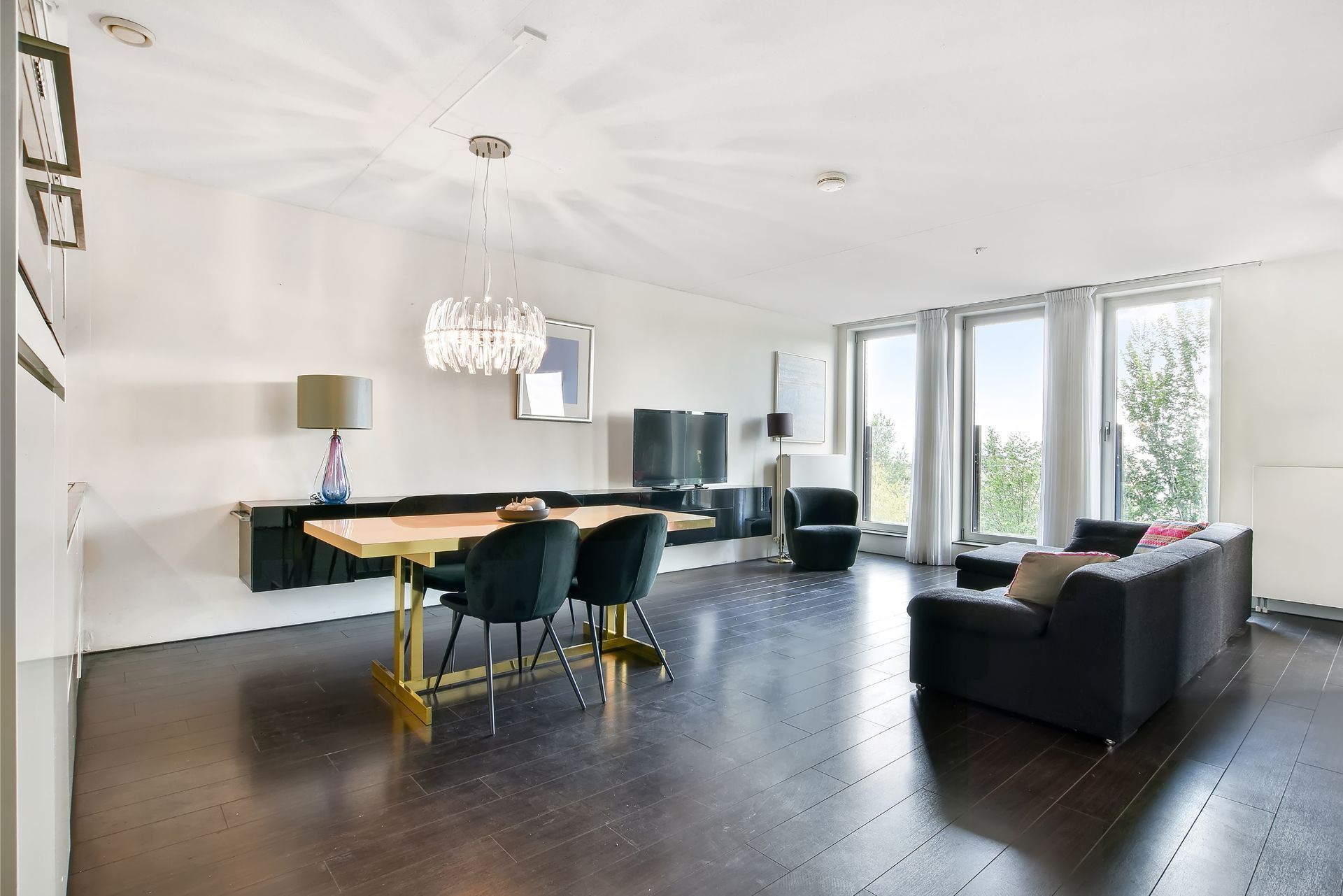 Modern open-plan living room with dark floors, dining table, sofa, and large windows with bright natural light