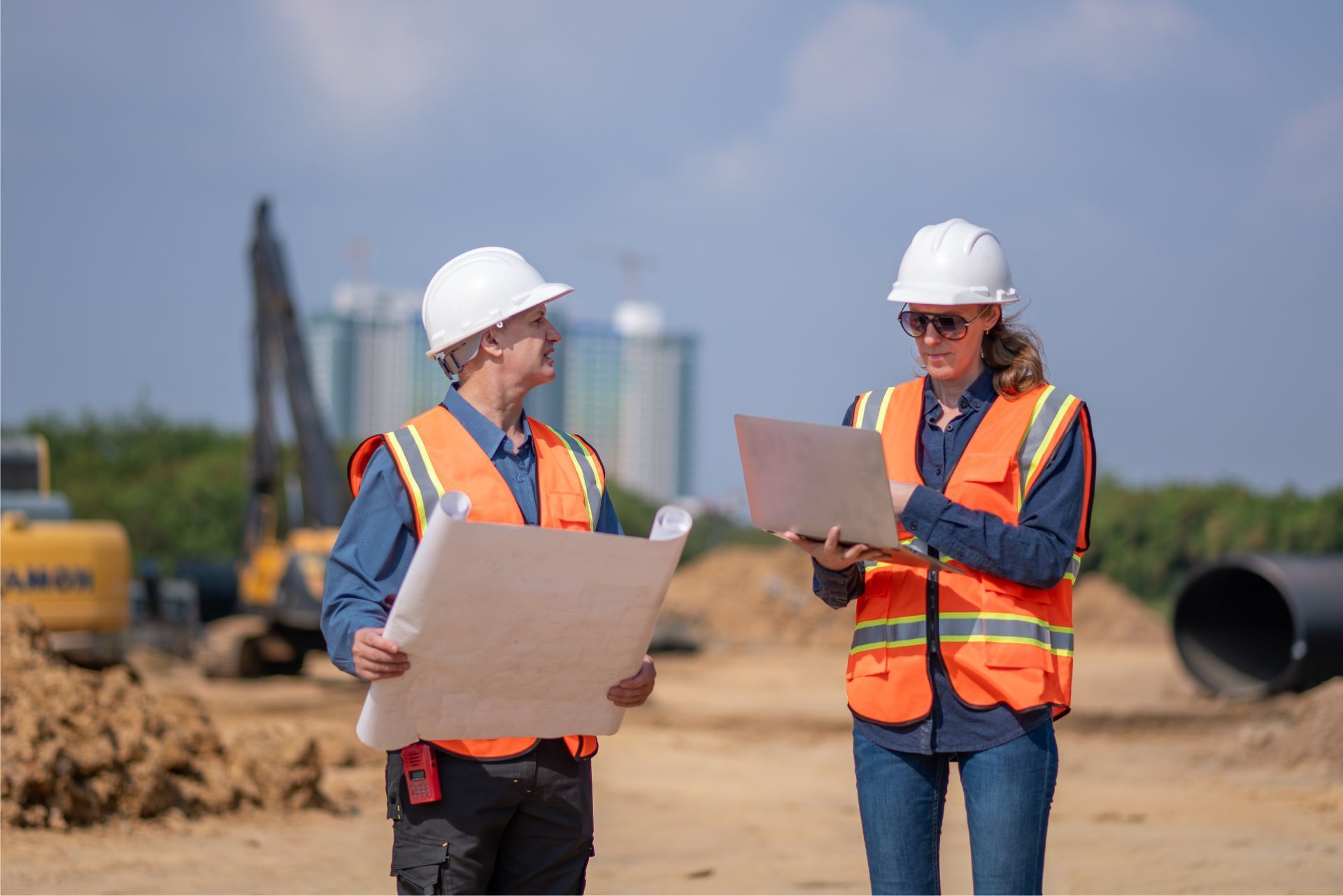 Two construction workers in hard hats review plans at a road construction site.