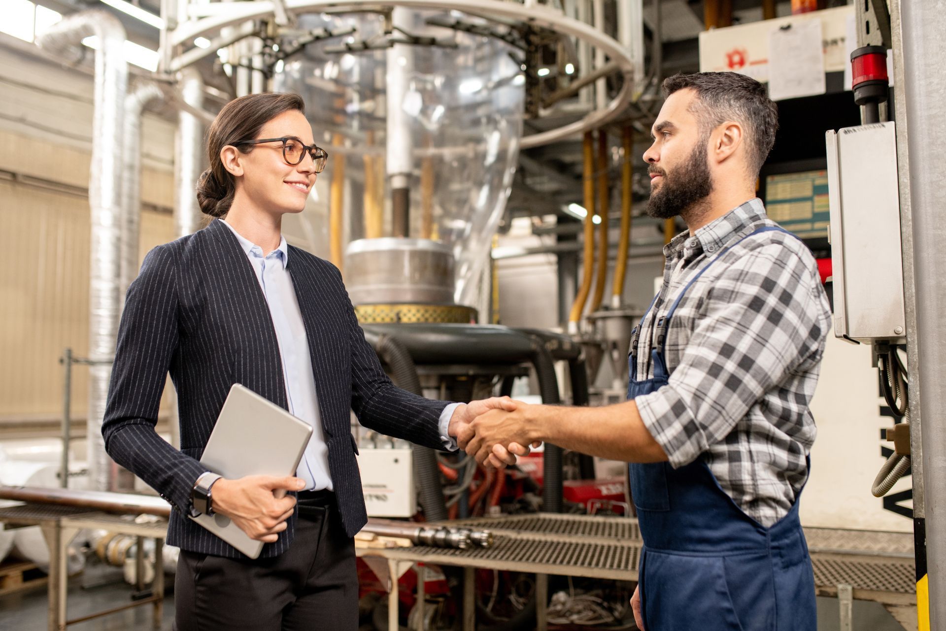 A professional in business attire shakes hands with a worker in uniform inside an industrial factory setting.