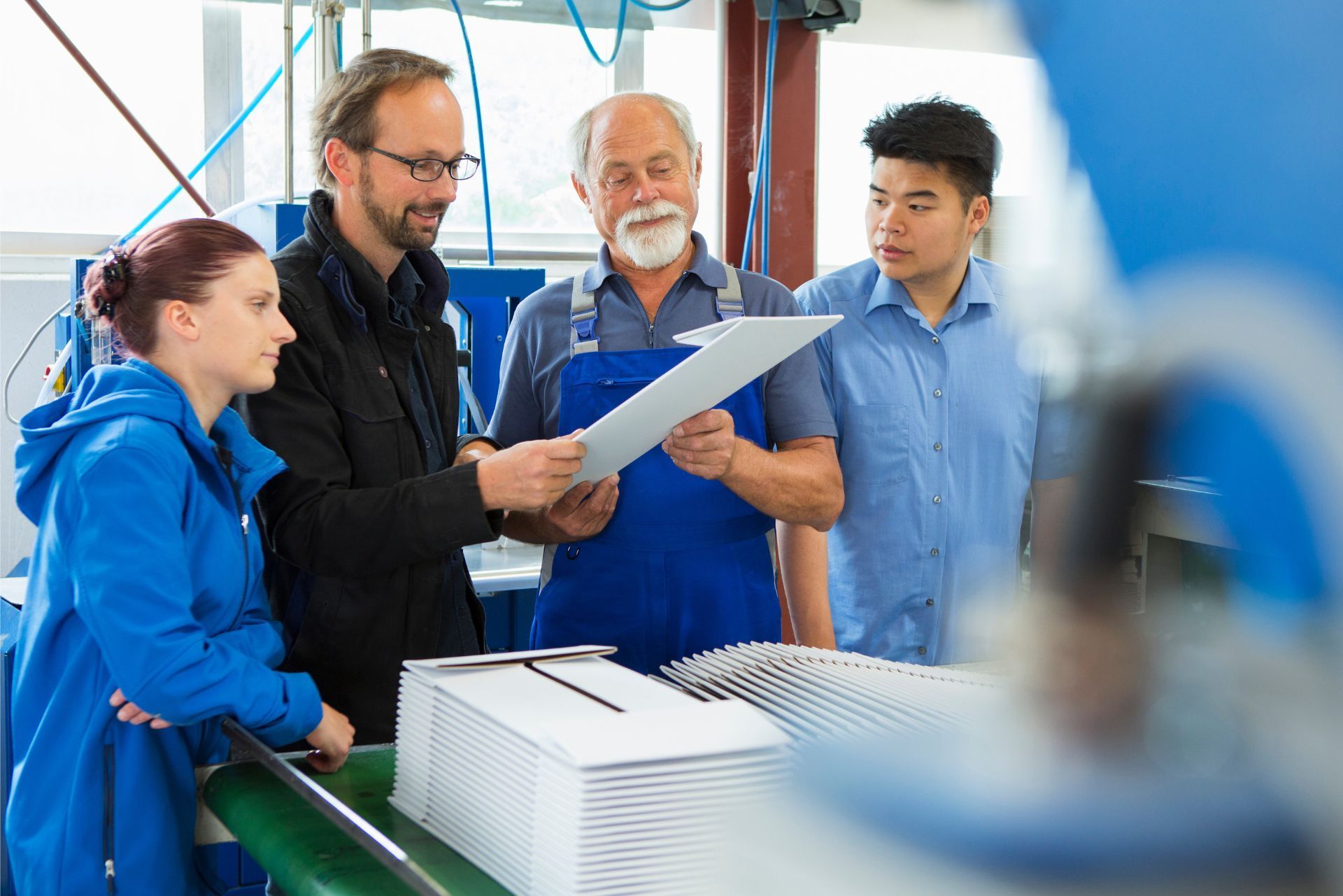 A group of people standing around a manufacturing table, inspecting a document together in a workshop environment.