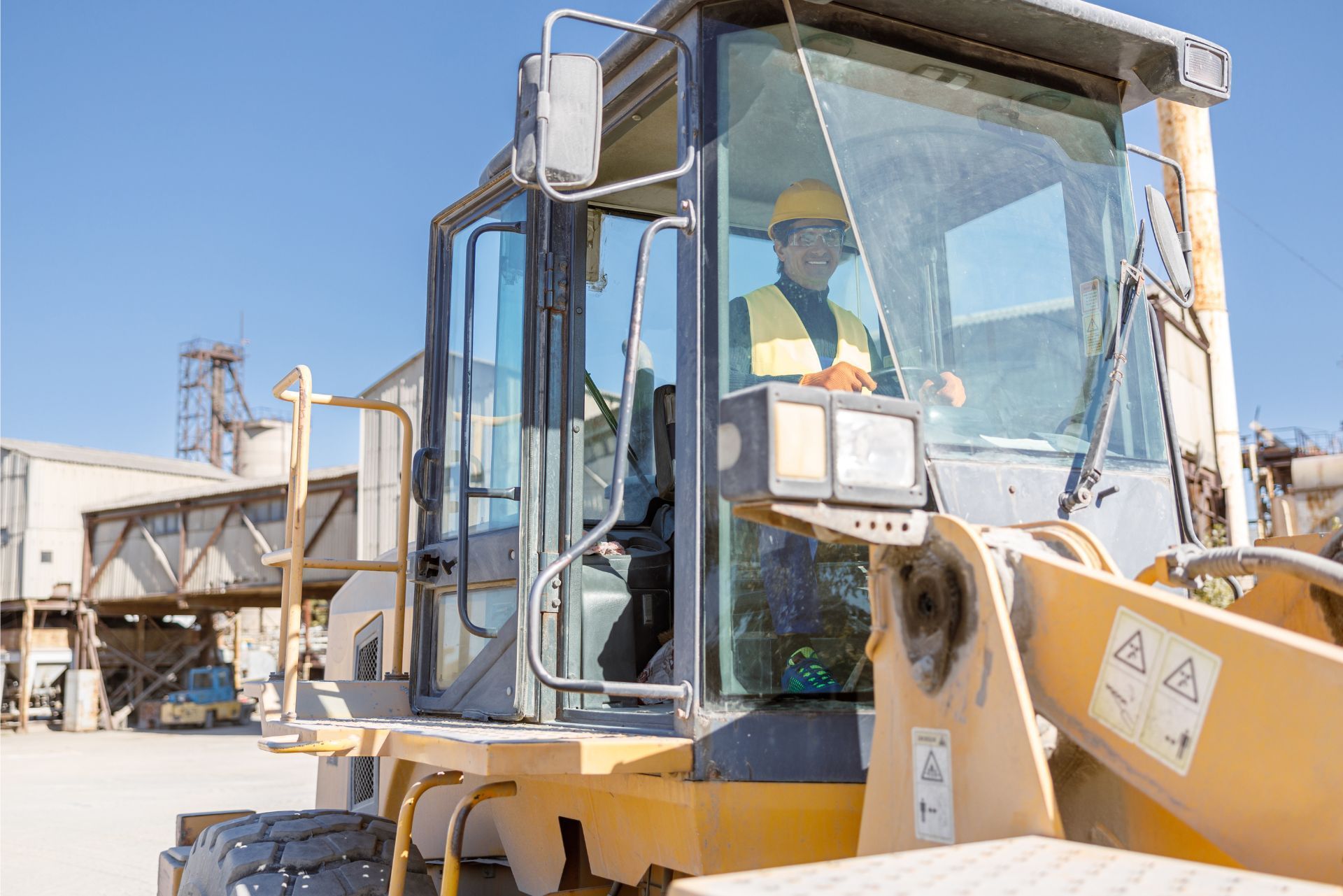 A worker wearing a yellow hard hat and safety vest operates a yellow construction loader at an industrial site.