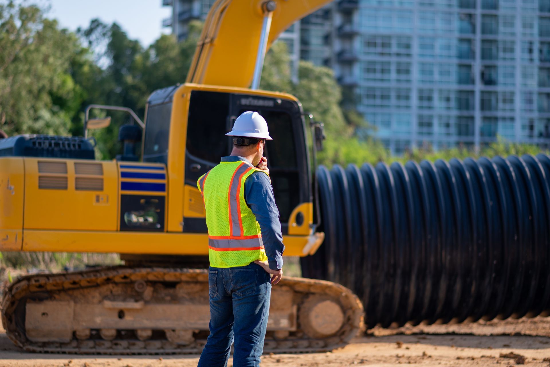 A construction worker in a high-visibility vest and hard hat stands before a yellow excavator and large black pipes.