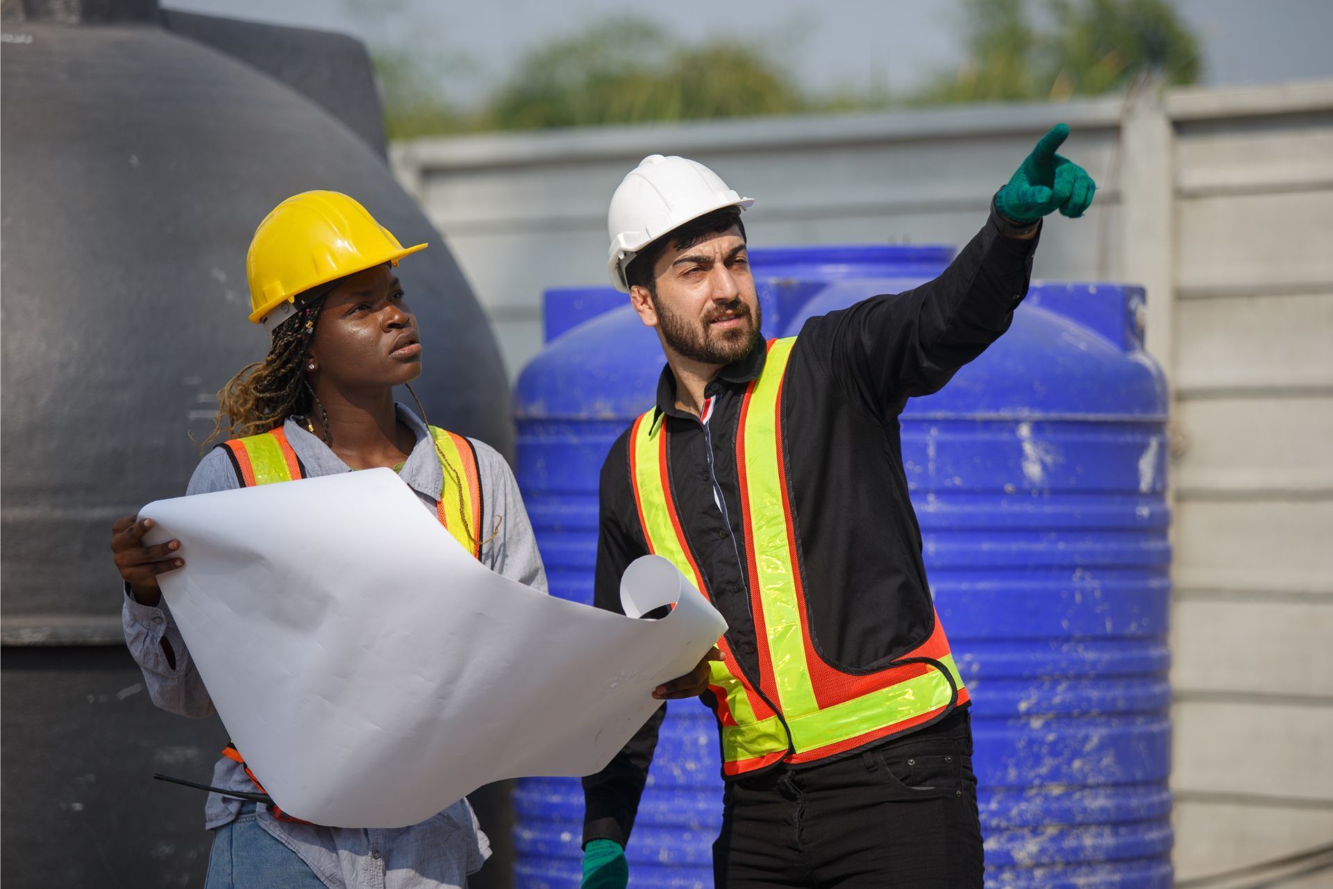 Two professionals in hard hats and safety vests stand outdoors reviewing blueprints as one points toward a work site.