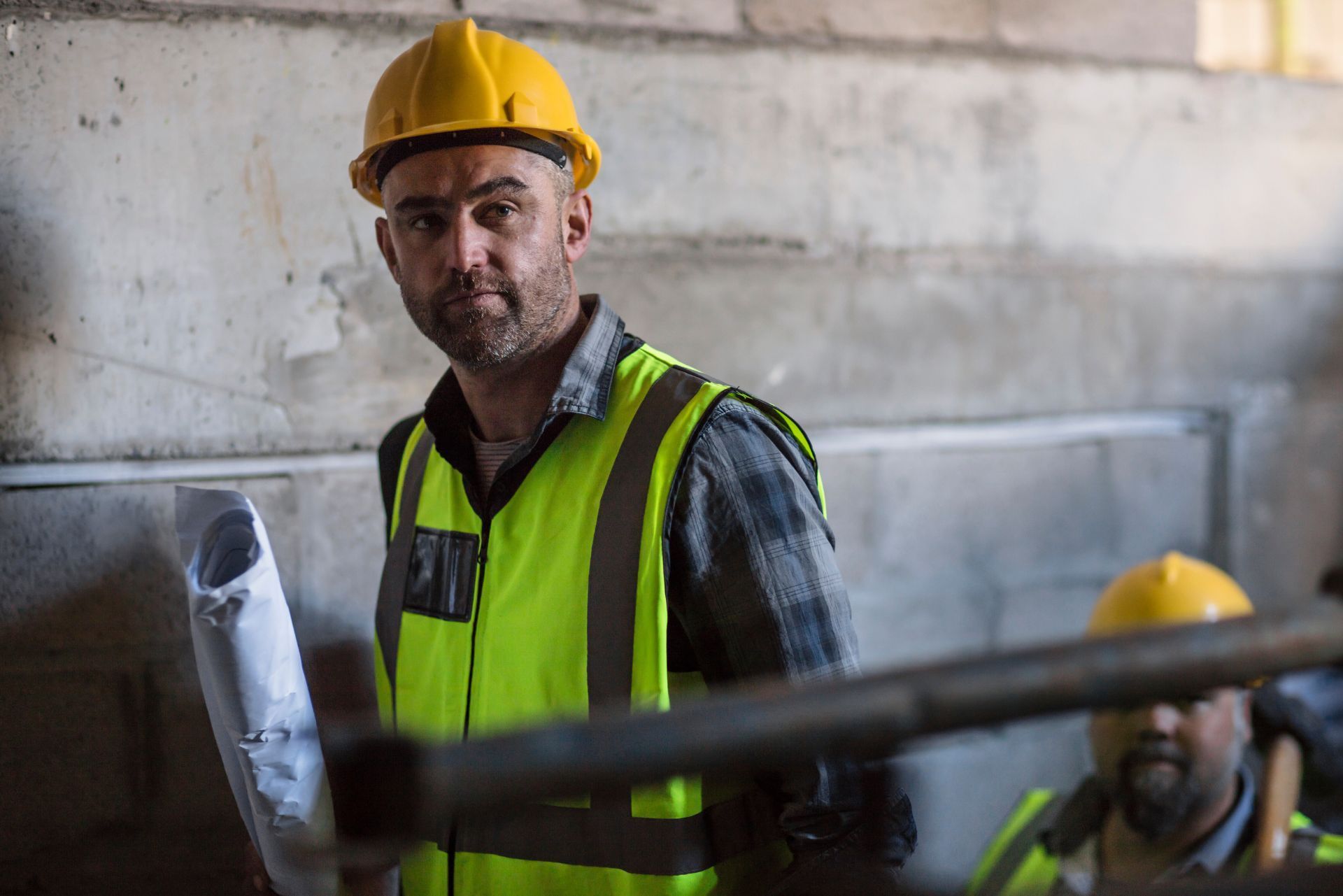 A construction worker in a yellow hard hat and high-visibility vest holds blueprints on a site, with a colleague behind.
