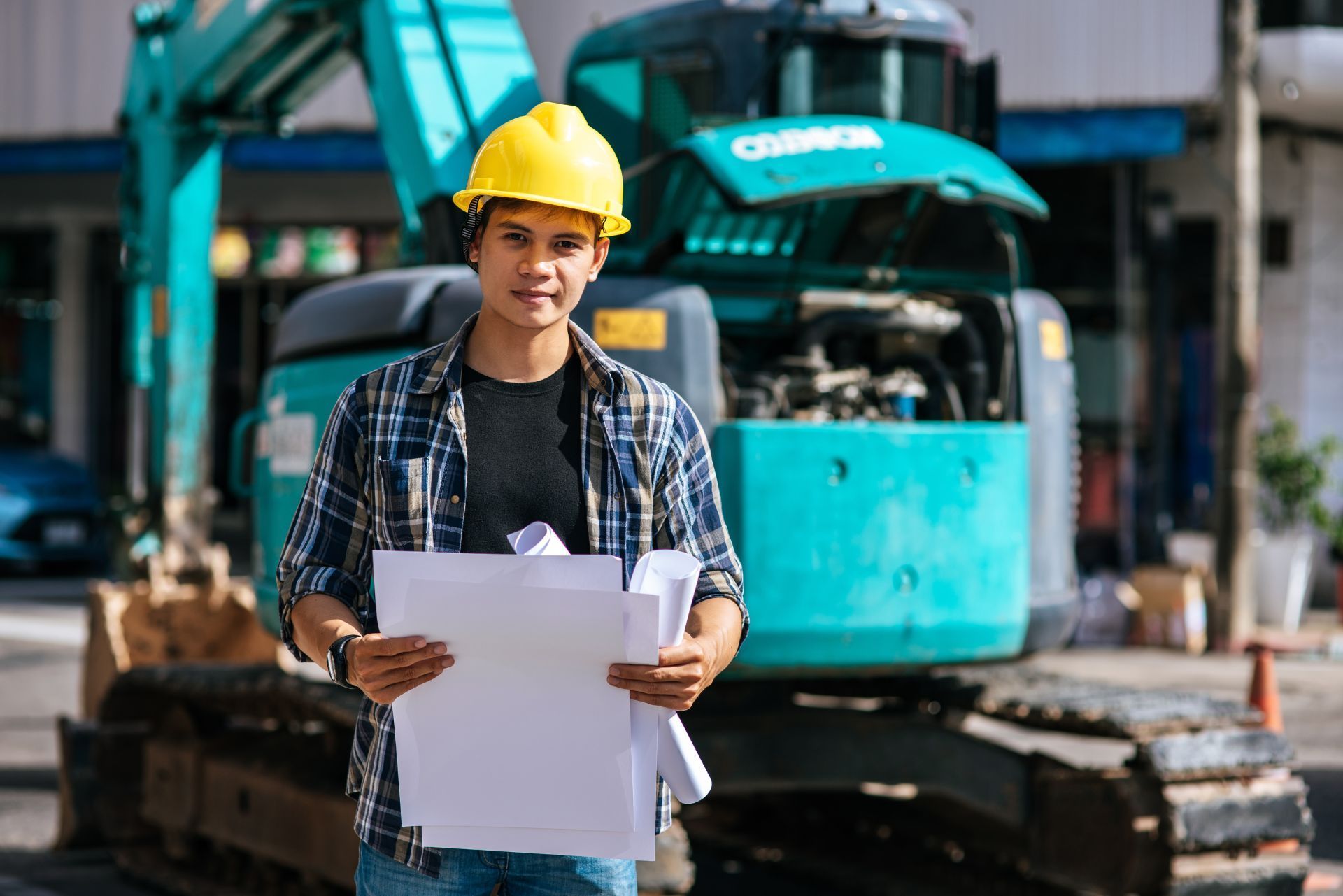 Construction worker in yellow hard hat holding blueprints in front of a turquoise excavator
